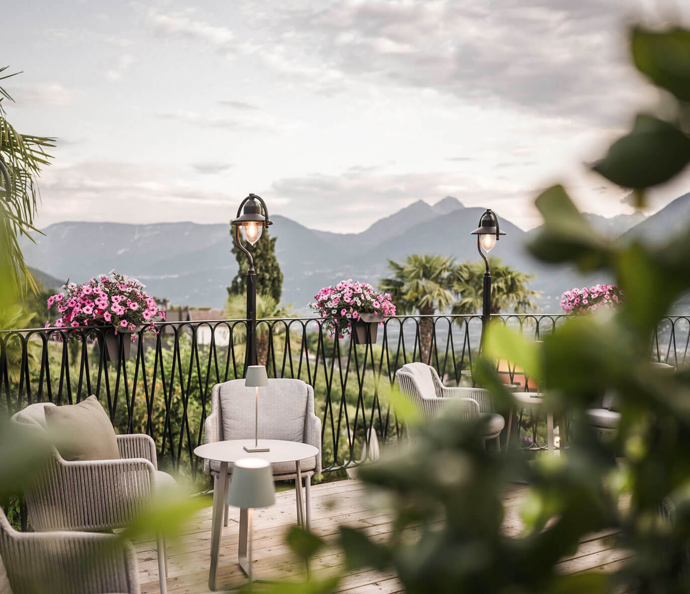 Chairs and tables on a scenic terrace with flowers, mountain views, and lamp posts at sunset. - Hotel Lagrein