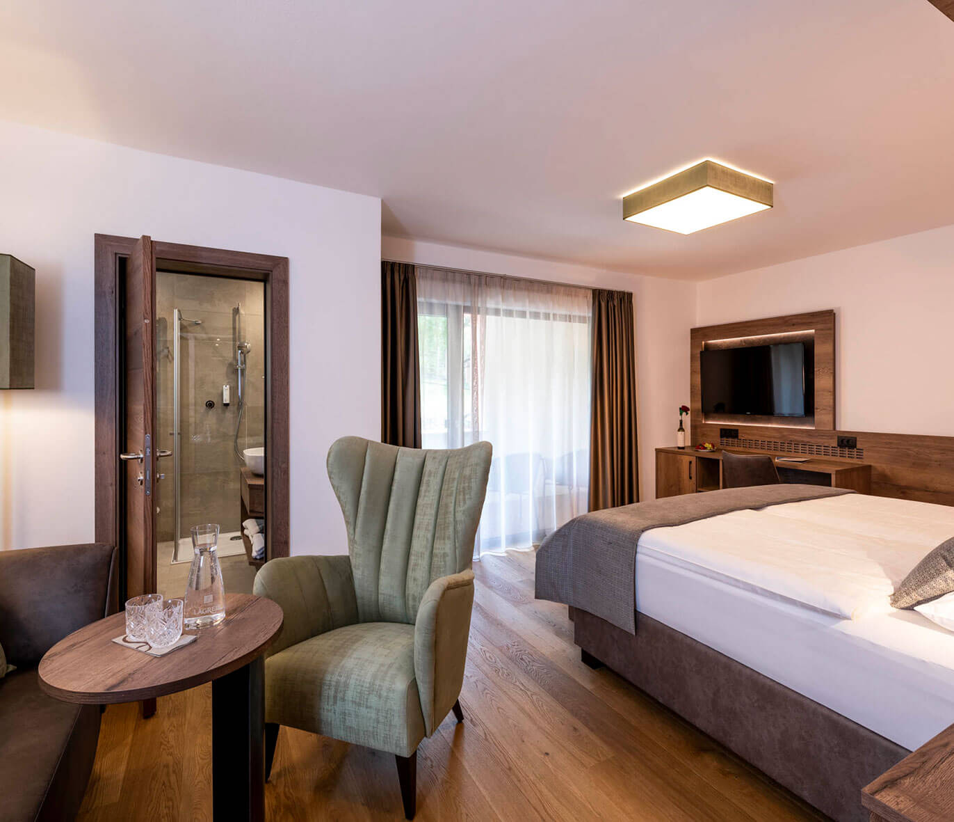 Modern hotel room with a double bed, armchair, TV, and private bathroom, featuring wood accents and natural light. - Hotel Lagrein