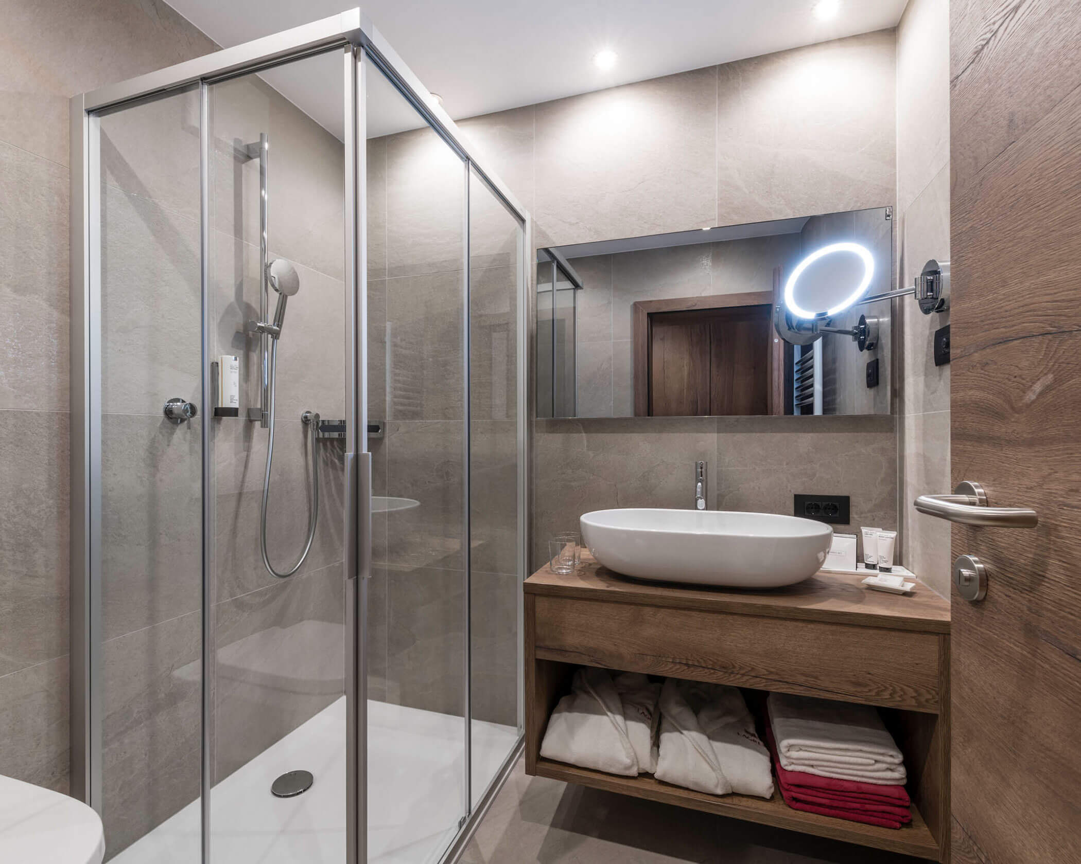 Modern bathroom with glass shower, vessel sink, large mirror, and folded towels on wooden shelves. - Hotel Lagrein