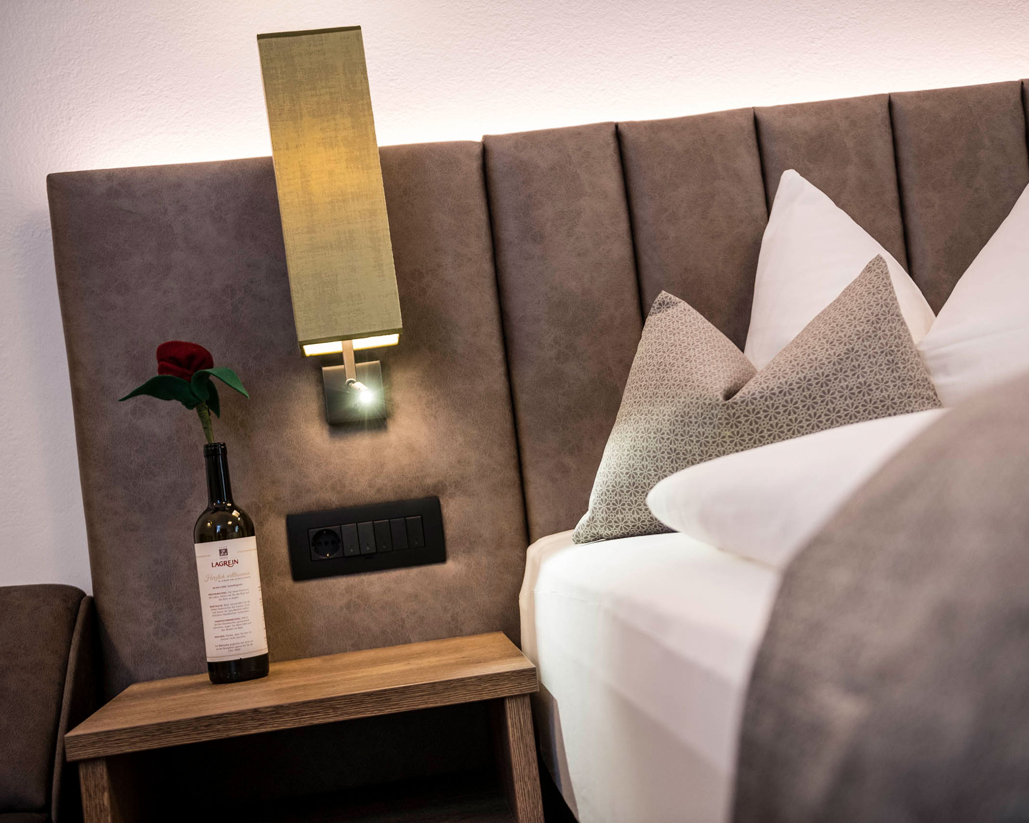 Bedside table with a wine bottle and rose, lamp above, and pillows arranged on a neatly made bed. - Hotel Lagrein