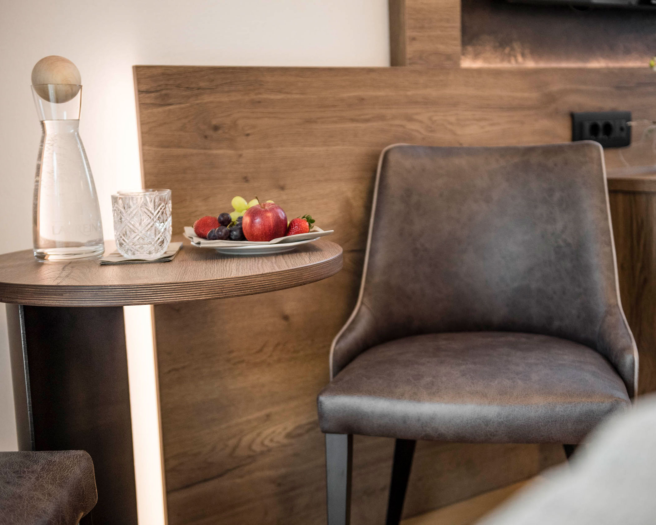 A glass carafe, a tumbler, and a fruit plate sit on a small table next to a brown chair and wooden wall. - Hotel Lagrein