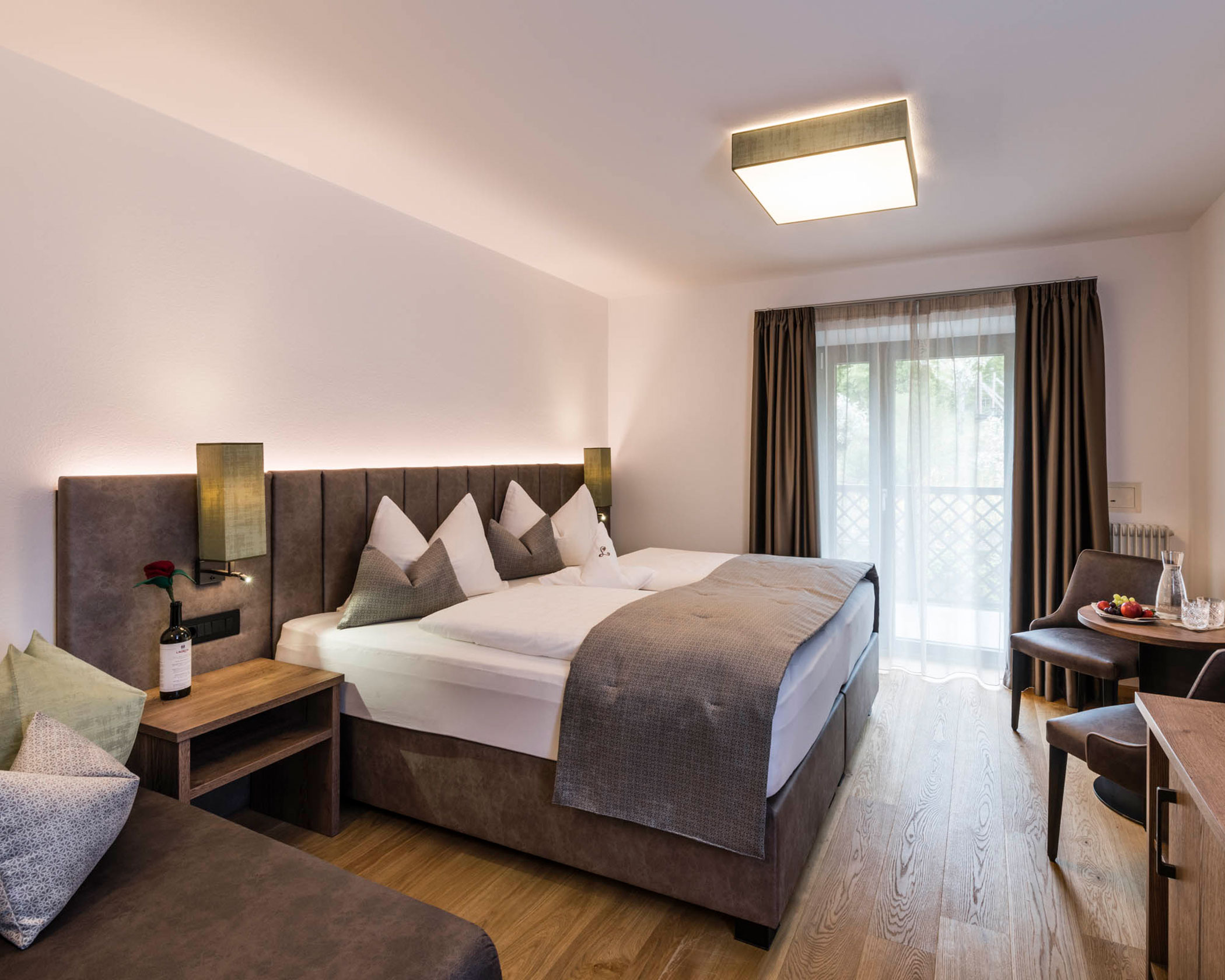 Modern hotel room with a large bed, seating area, wooden floor, and large window with curtains. - Hotel Lagrein