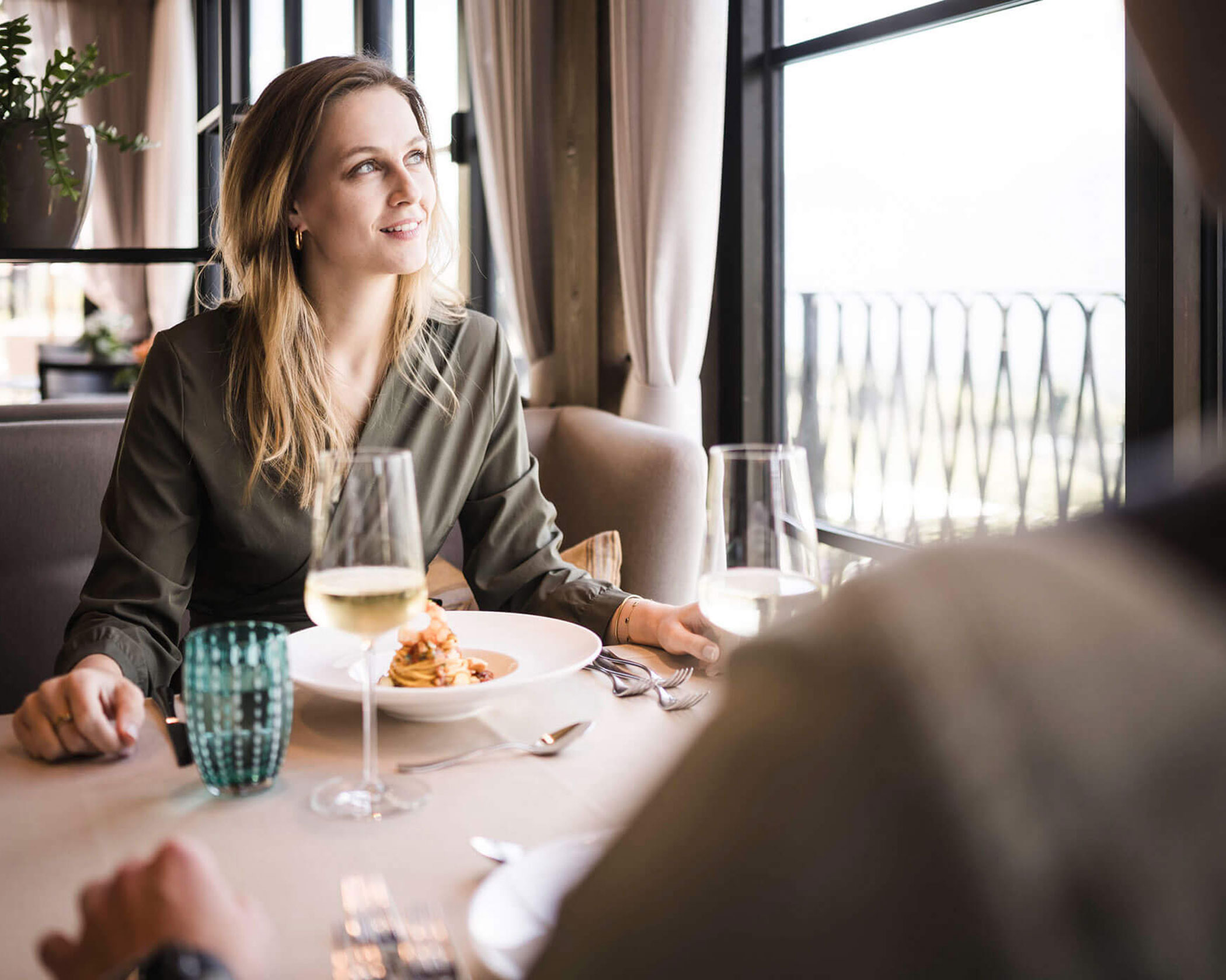 A woman sits at a restaurant table by a window, smiling with food and wine in front of her. - Hotel Lagrein