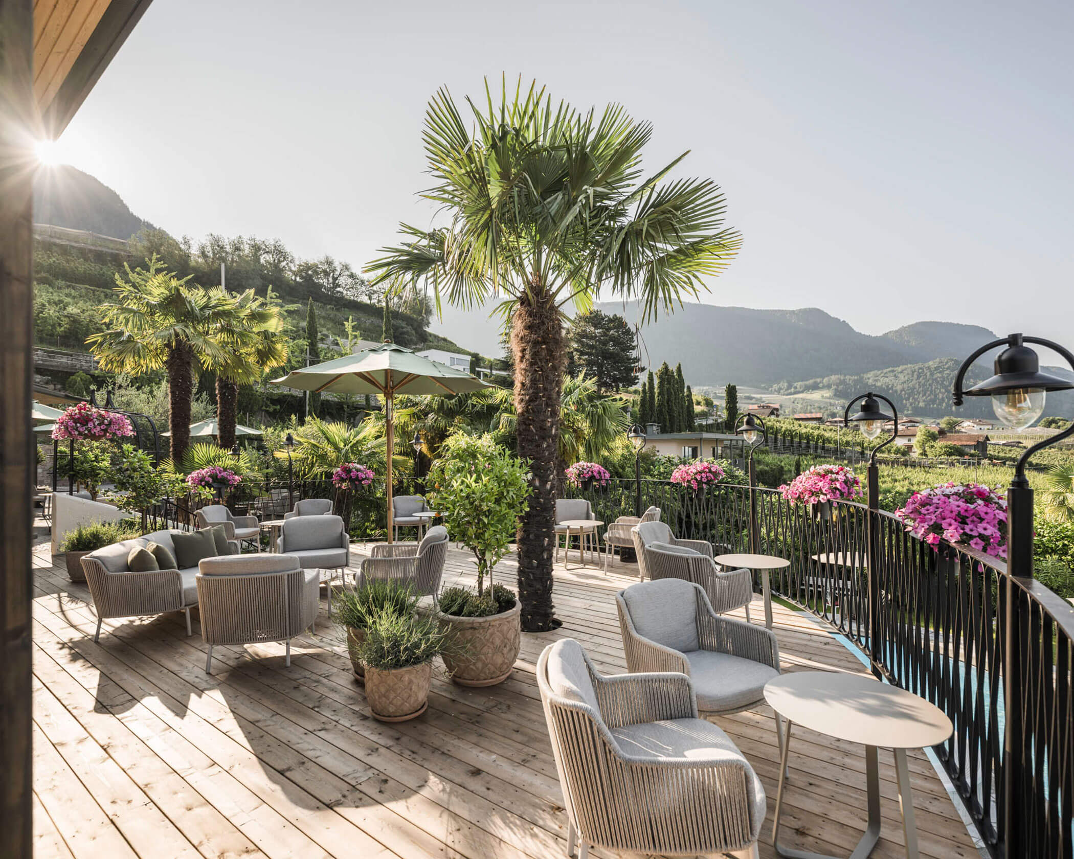 Outdoor terrace with palm trees, gray lounge chairs, flowers, and mountain views under a clear sky. - Hotel Lagrein