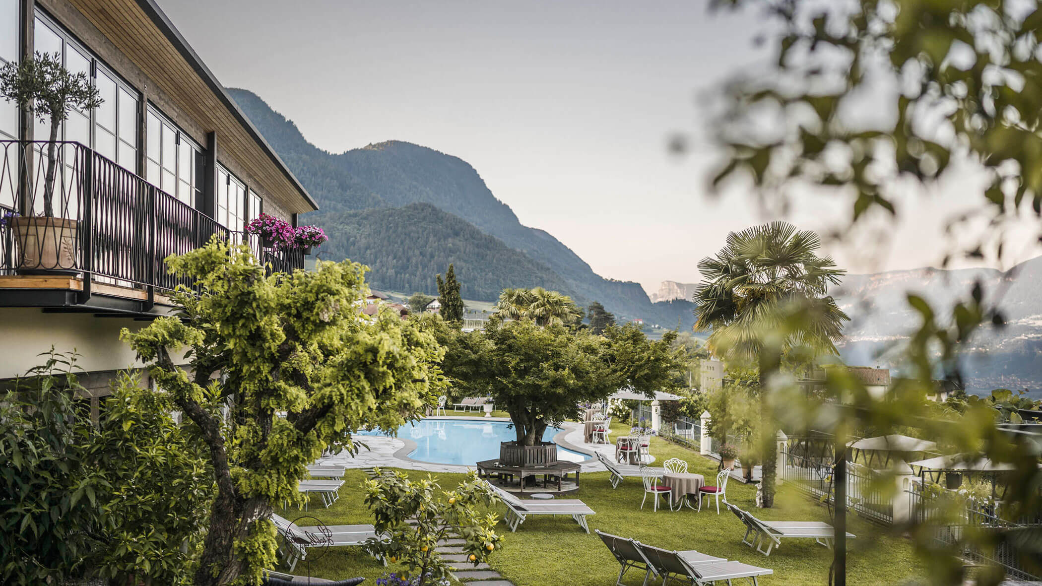 Hotel garden with pool, lounge chairs, trees, and mountain views in the background. - Hotel Lagrein