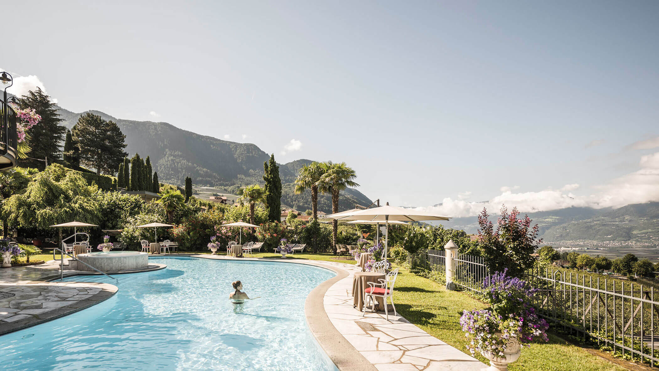 A person swims in a curved outdoor pool with mountain views, surrounded by lush greenery and umbrellas. - Hotel Lagrein
