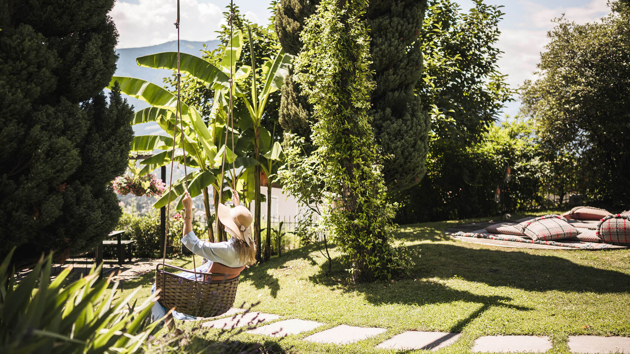 Person wearing a hat swings in a garden surrounded by lush greenery on a sunny day. - Hotel Lagrein