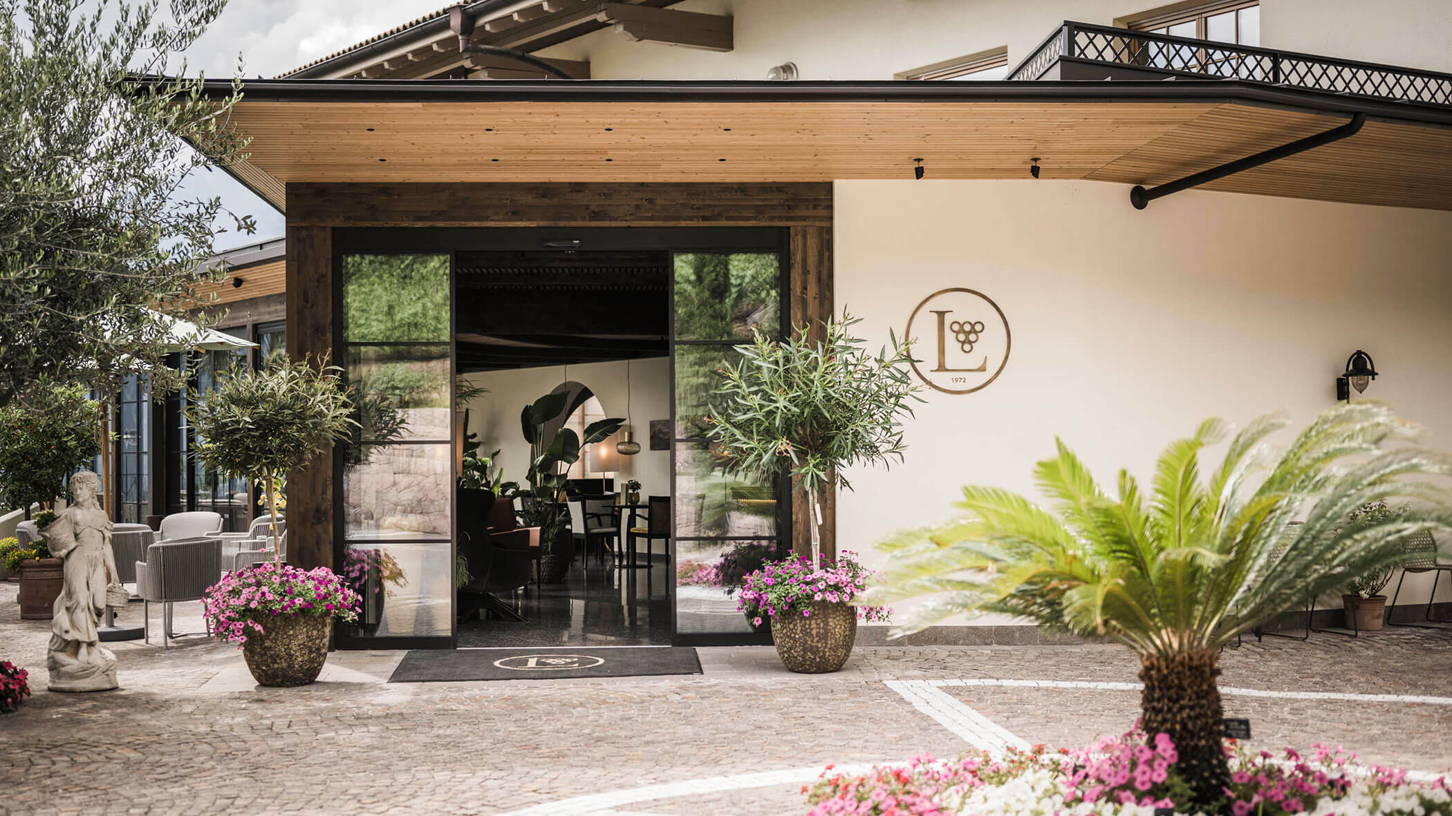 Modern building entrance with glass doors, potted plants, and flowers on a cobblestone pathway. - Hotel Lagrein