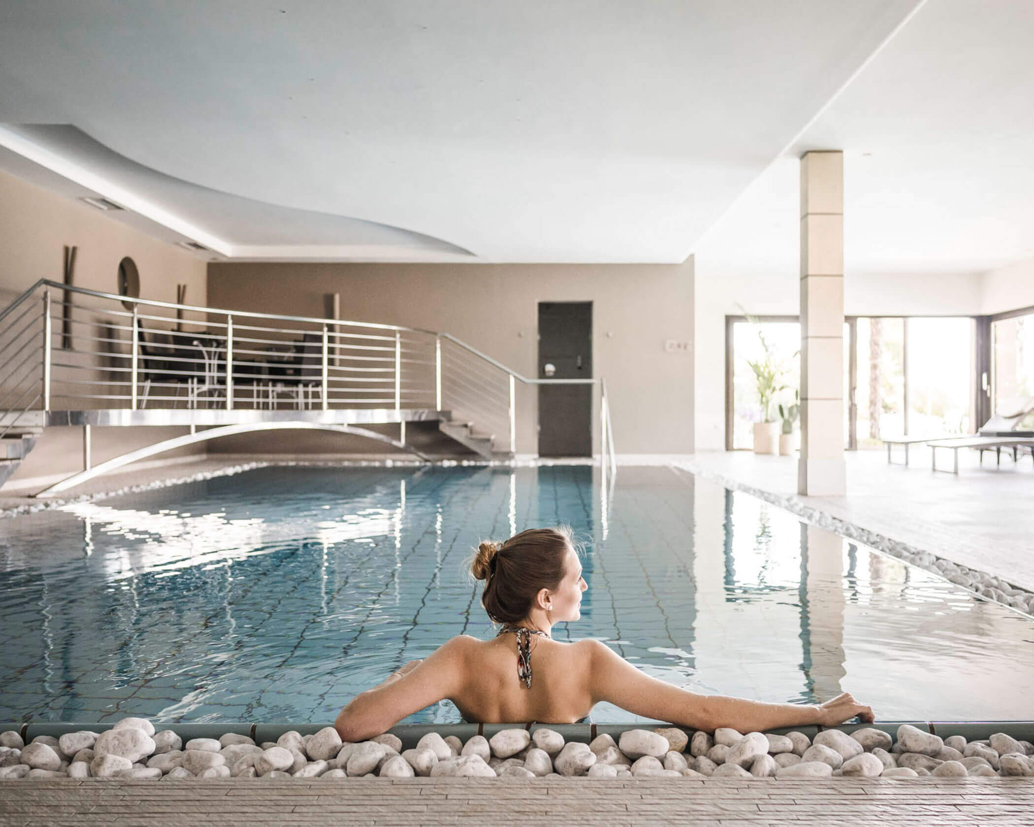 Woman relaxing in an indoor pool, leaning on the edge with a small bridge and lounge chairs in the background. - Hotel Lagrein