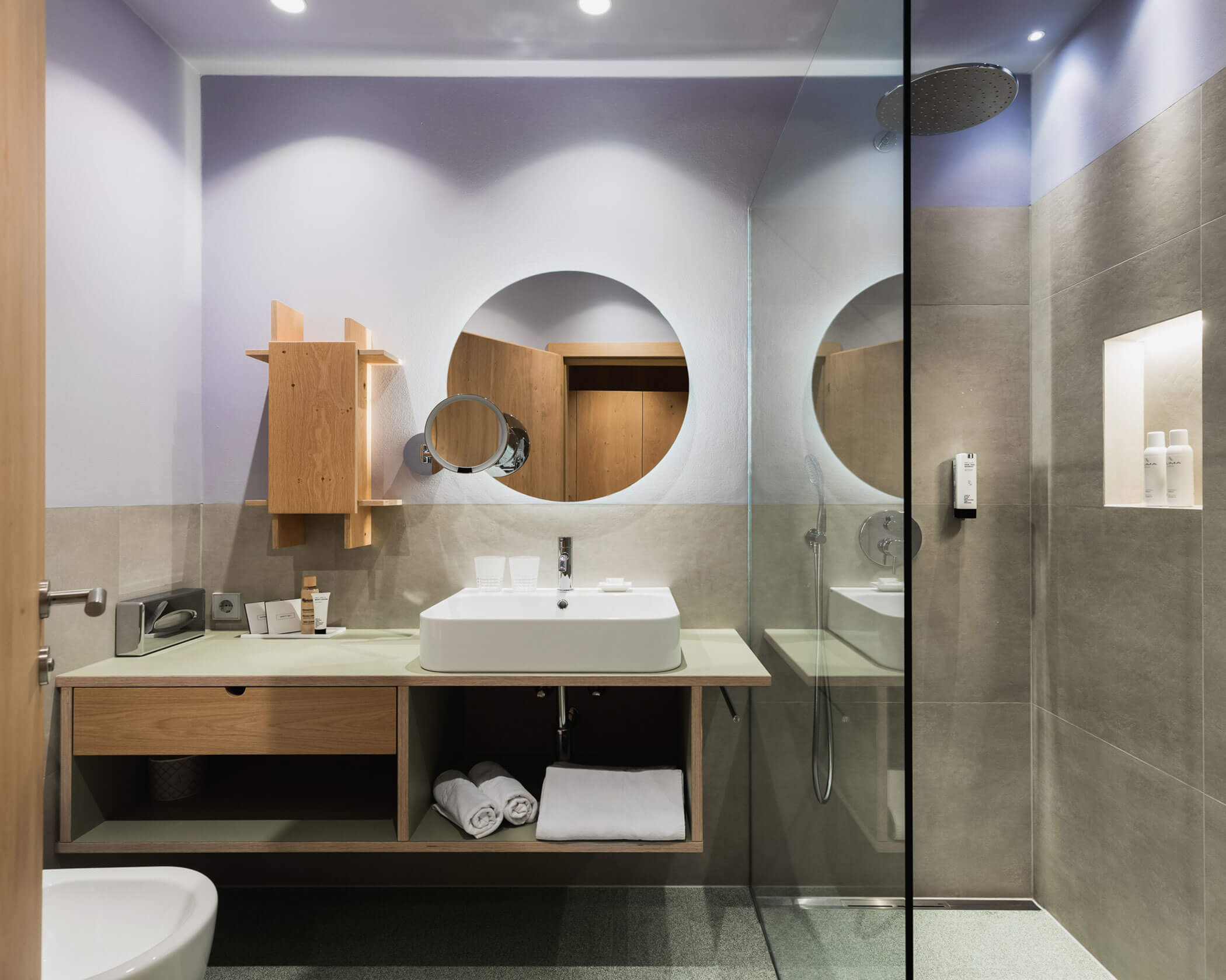 Modern bathroom with a round mirror, sink, wooden cabinets, and a glass-walled shower area. - Hotel Lagrein