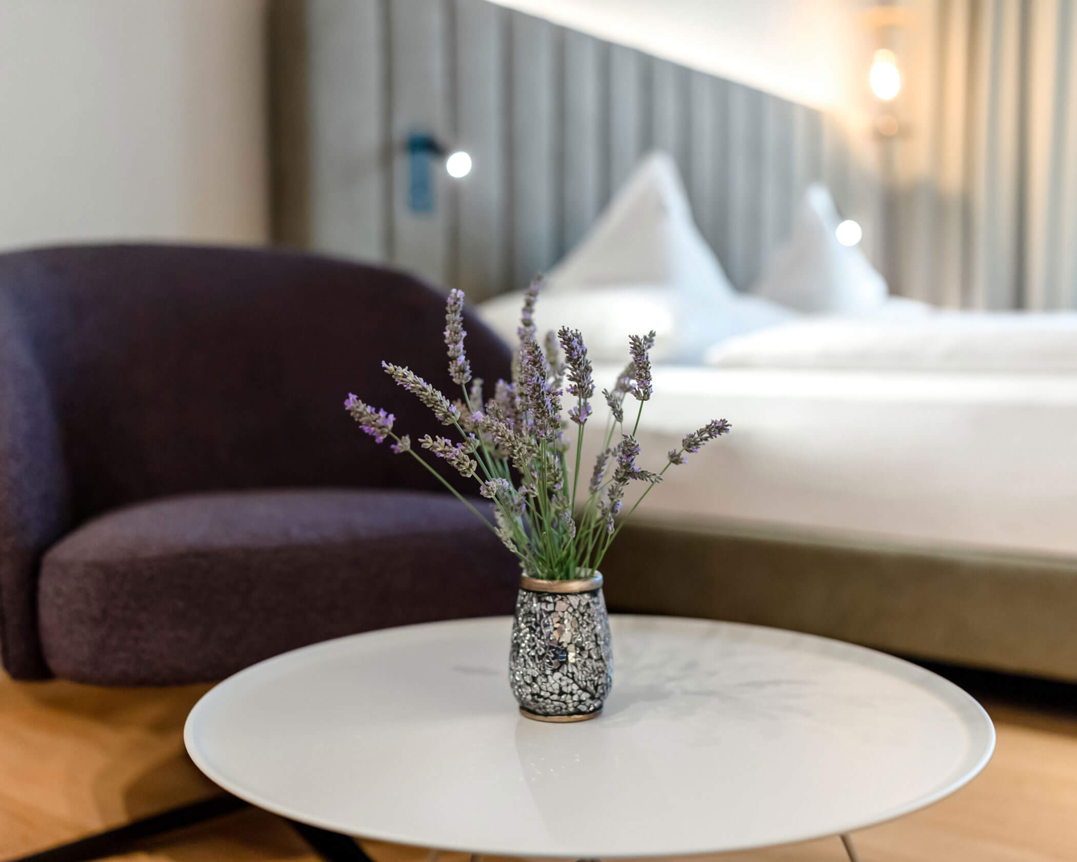 A small vase with lavender sits on a round white table in a modern bedroom with a bed and purple chair. - Hotel Lagrein