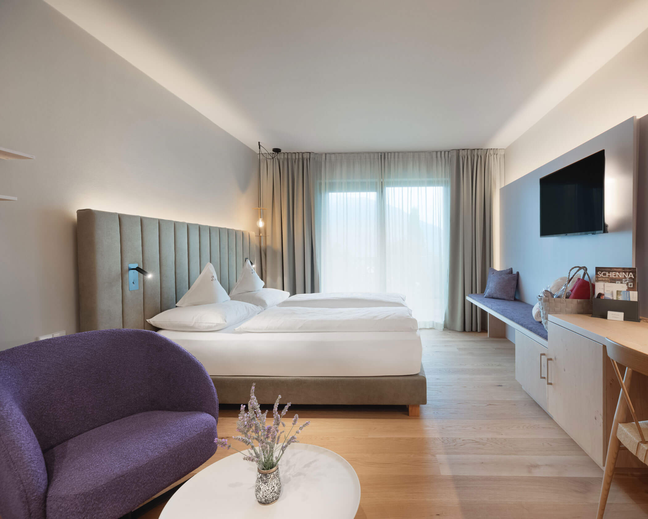 Modern hotel room with a double bed, purple chair, TV, and large window with sheer curtains. - Hotel Lagrein