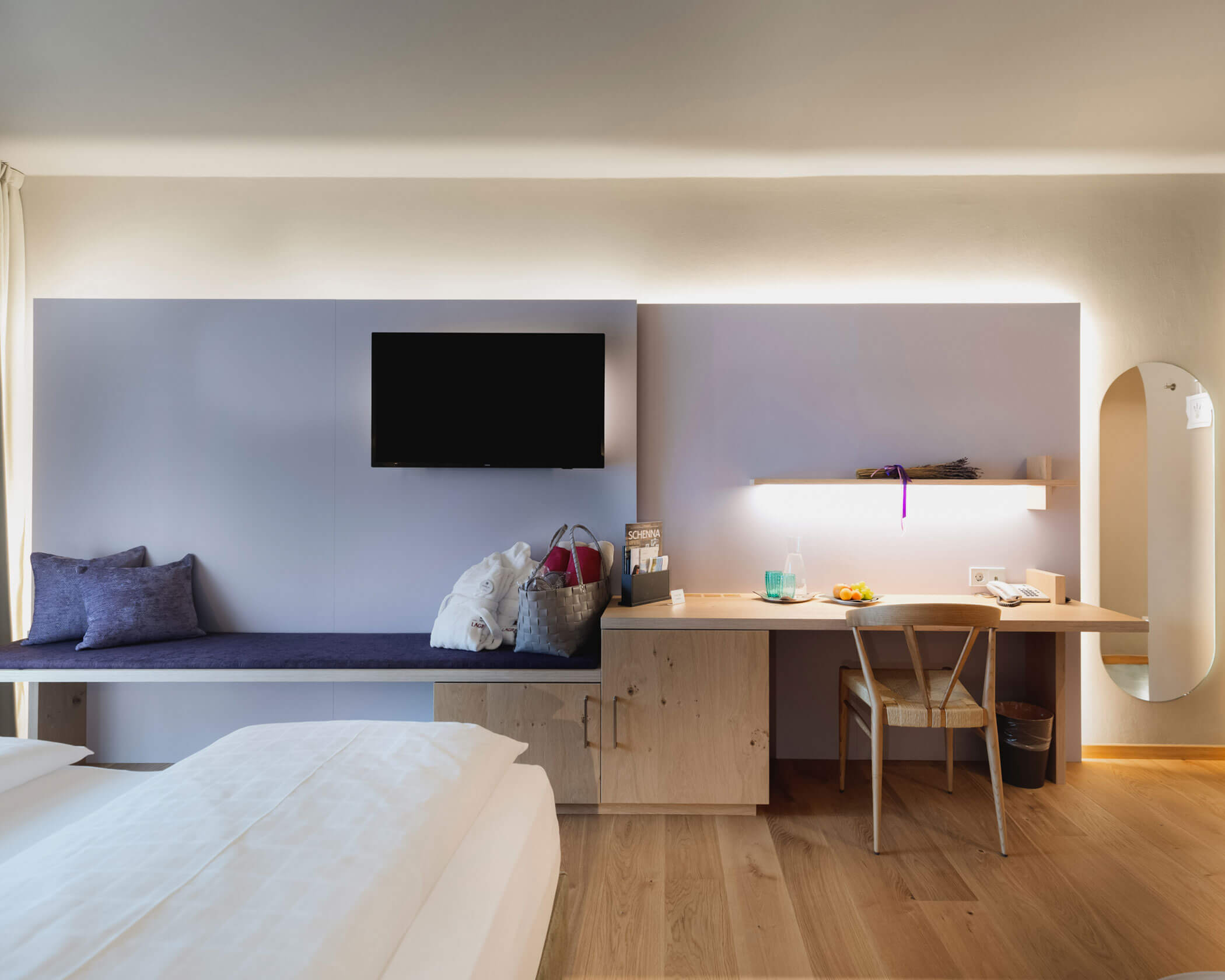 Modern hotel room with a bed, desk, wall-mounted TV, bench seating, and a mirror on a wooden floor. - Hotel Lagrein