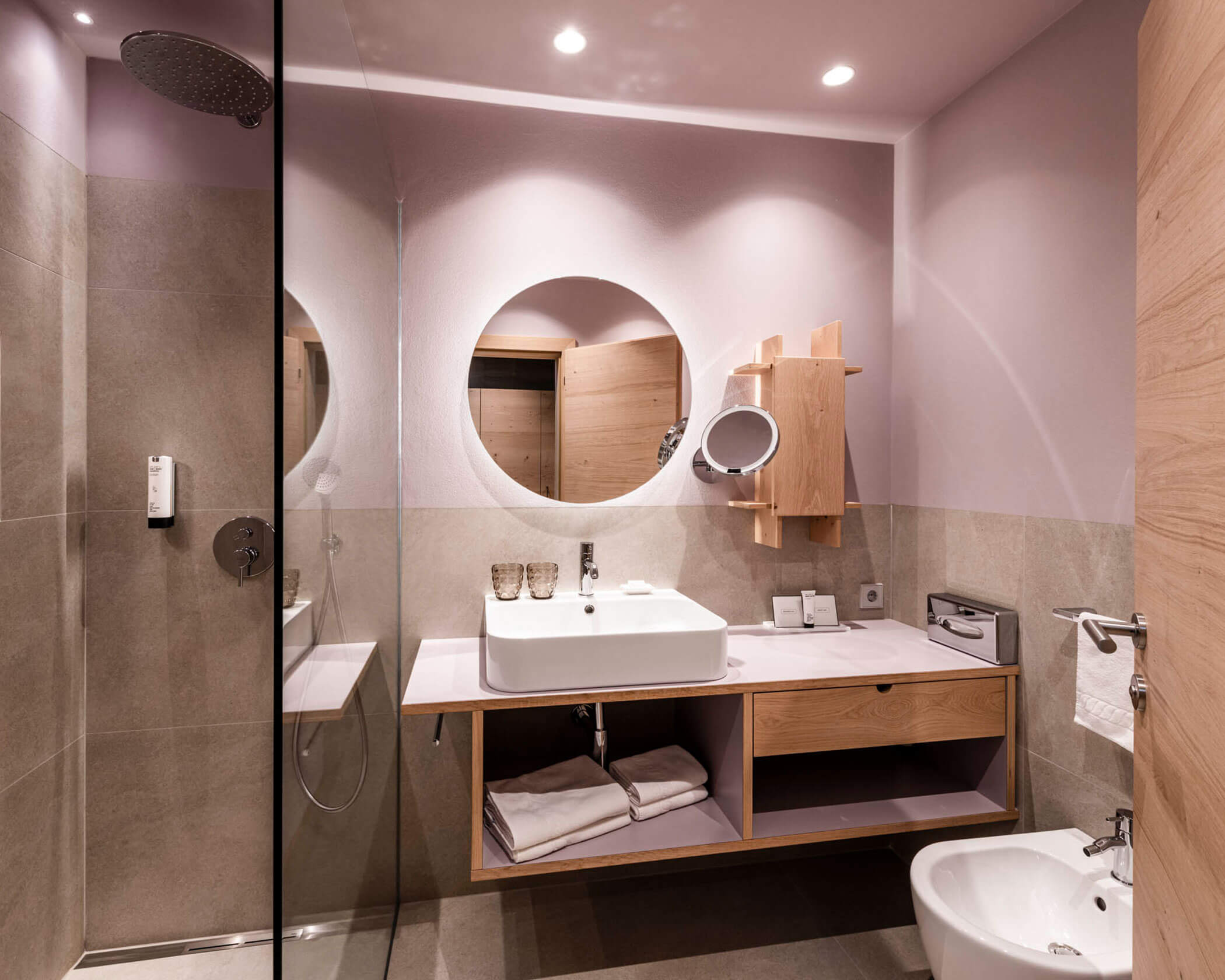 Modern bathroom with a round mirror, sink, shower, and bidet, featuring neutral colors and wood accents. - Hotel Lagrein