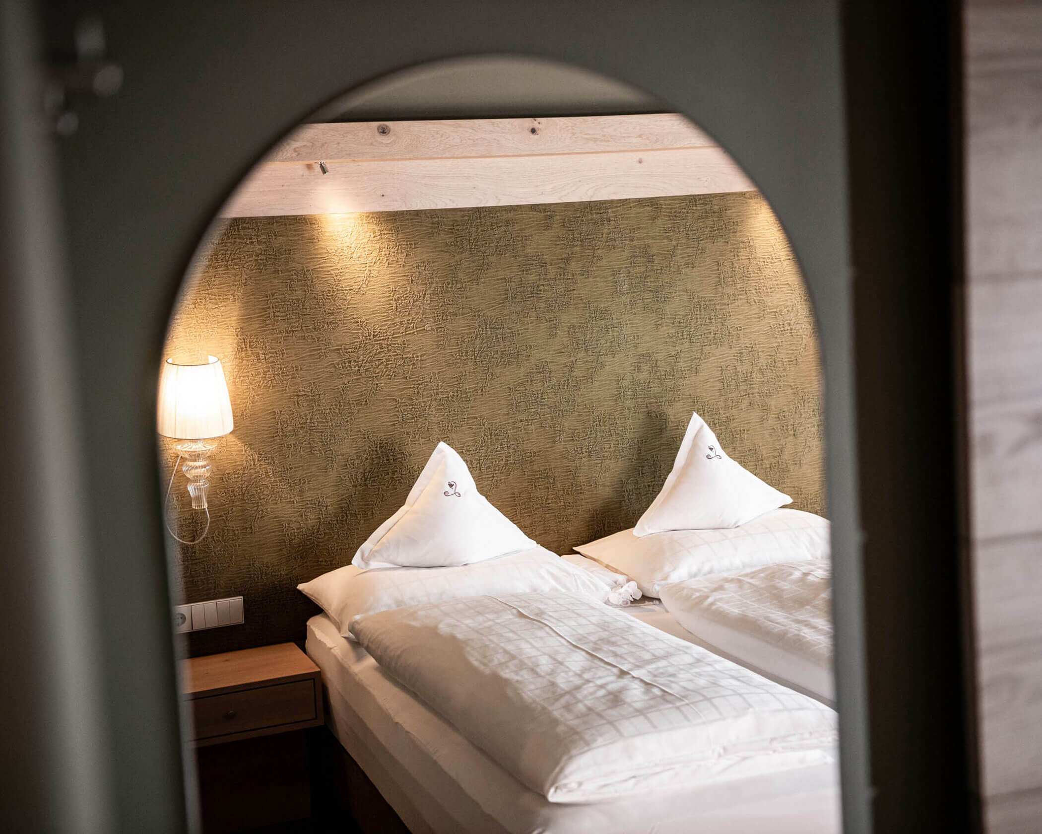 Two neatly made twin beds with white linens and pillows, viewed through an archway in a cozy room. - Hotel Lagrein