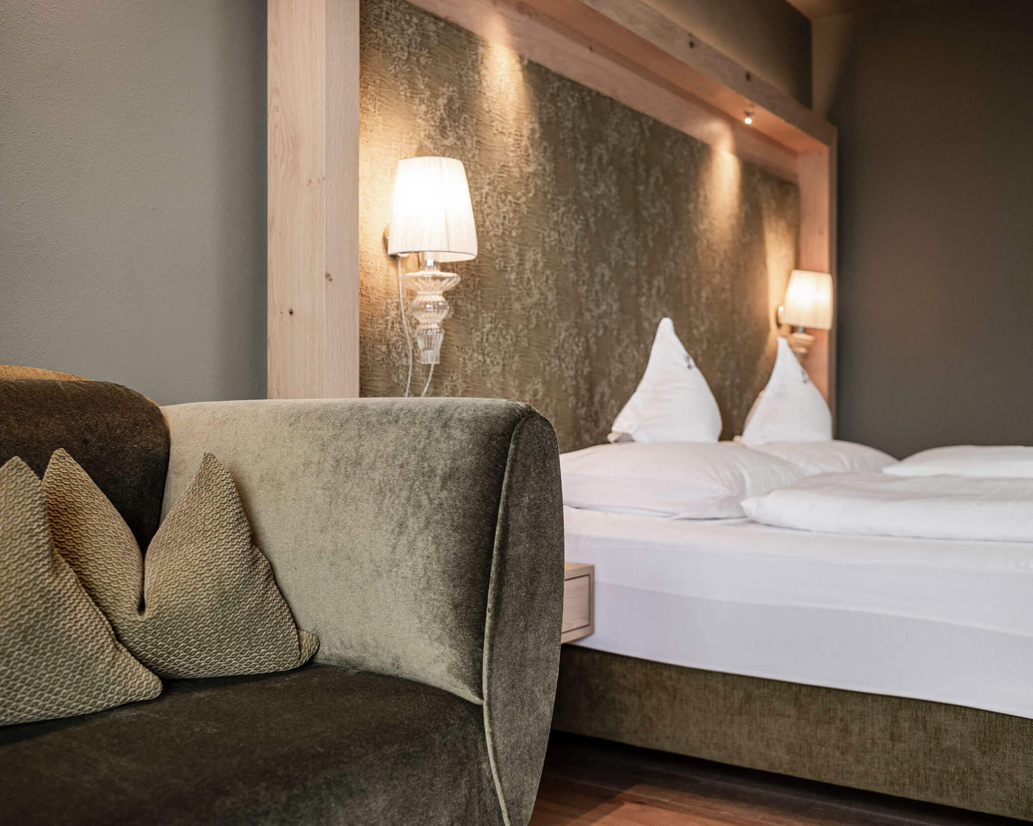 A modern hotel room with a green sofa, decorative pillows, and a double bed with white linens and wall lamps. - Hotel Lagrein