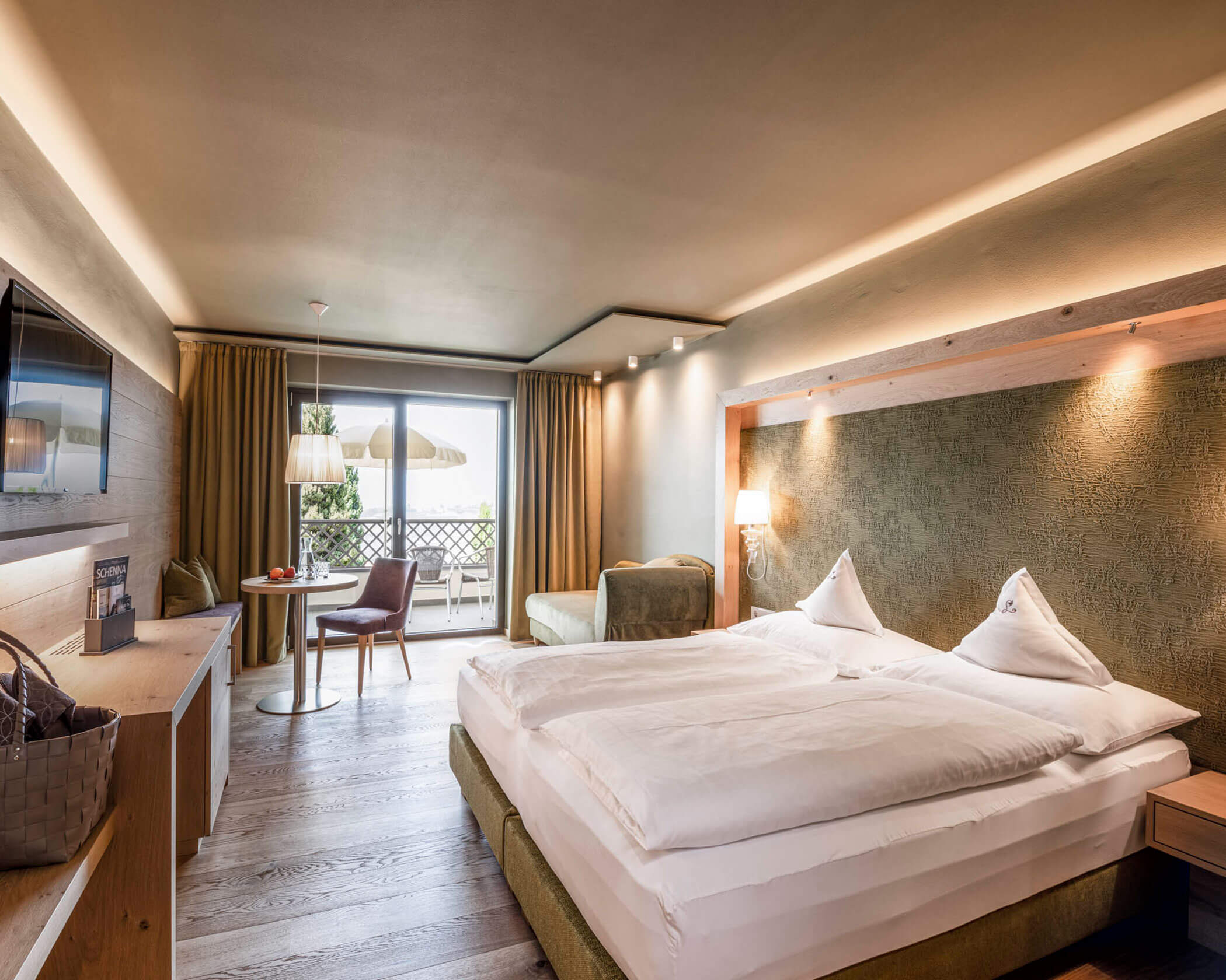 Bright hotel room with double bed, sofa, TV, desk, and balcony with outdoor seating and natural light. - Hotel Lagrein