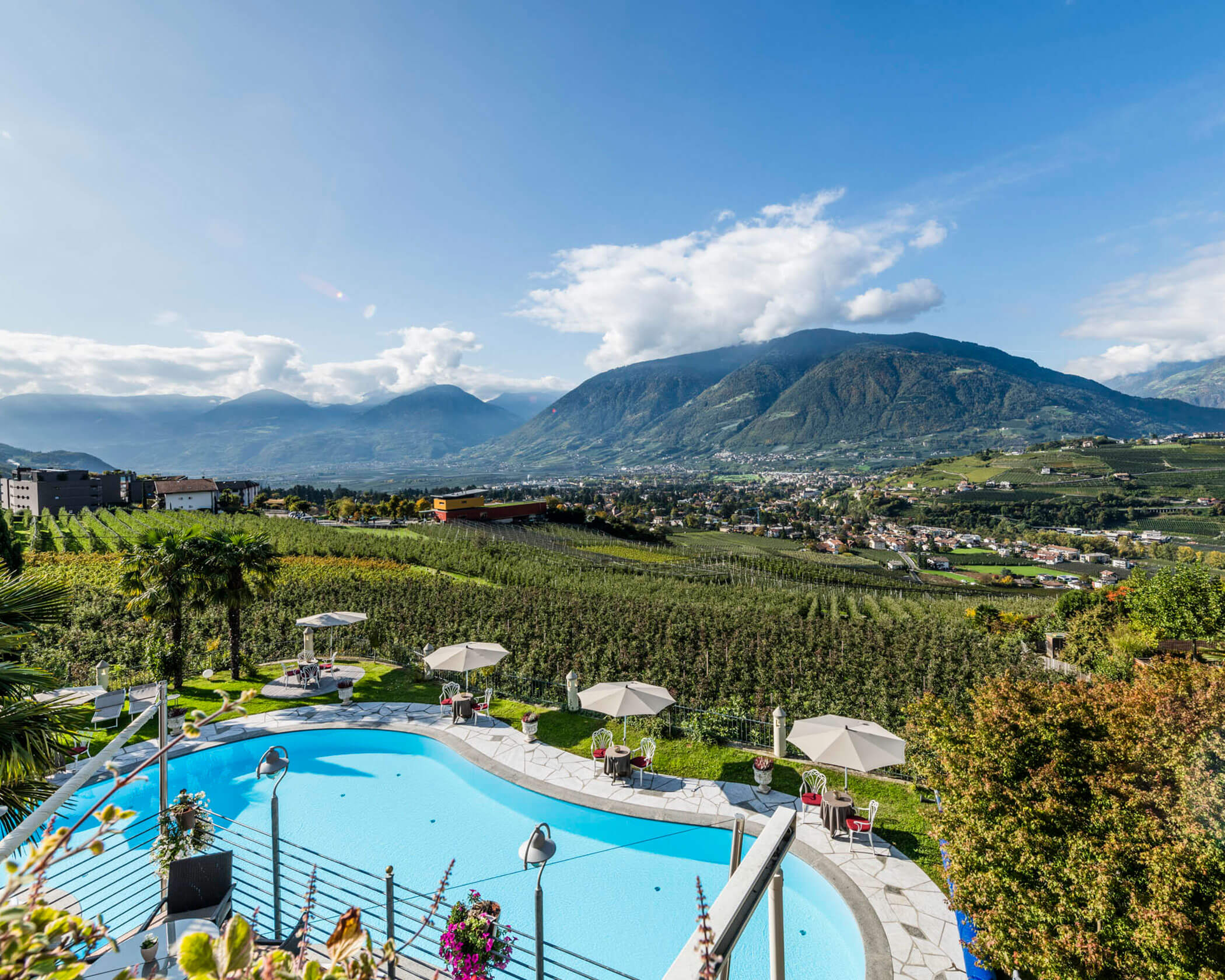 Outdoor pool with umbrellas overlooking vineyards, a valley, and mountains under a blue sky with some clouds. - Hotel Lagrein