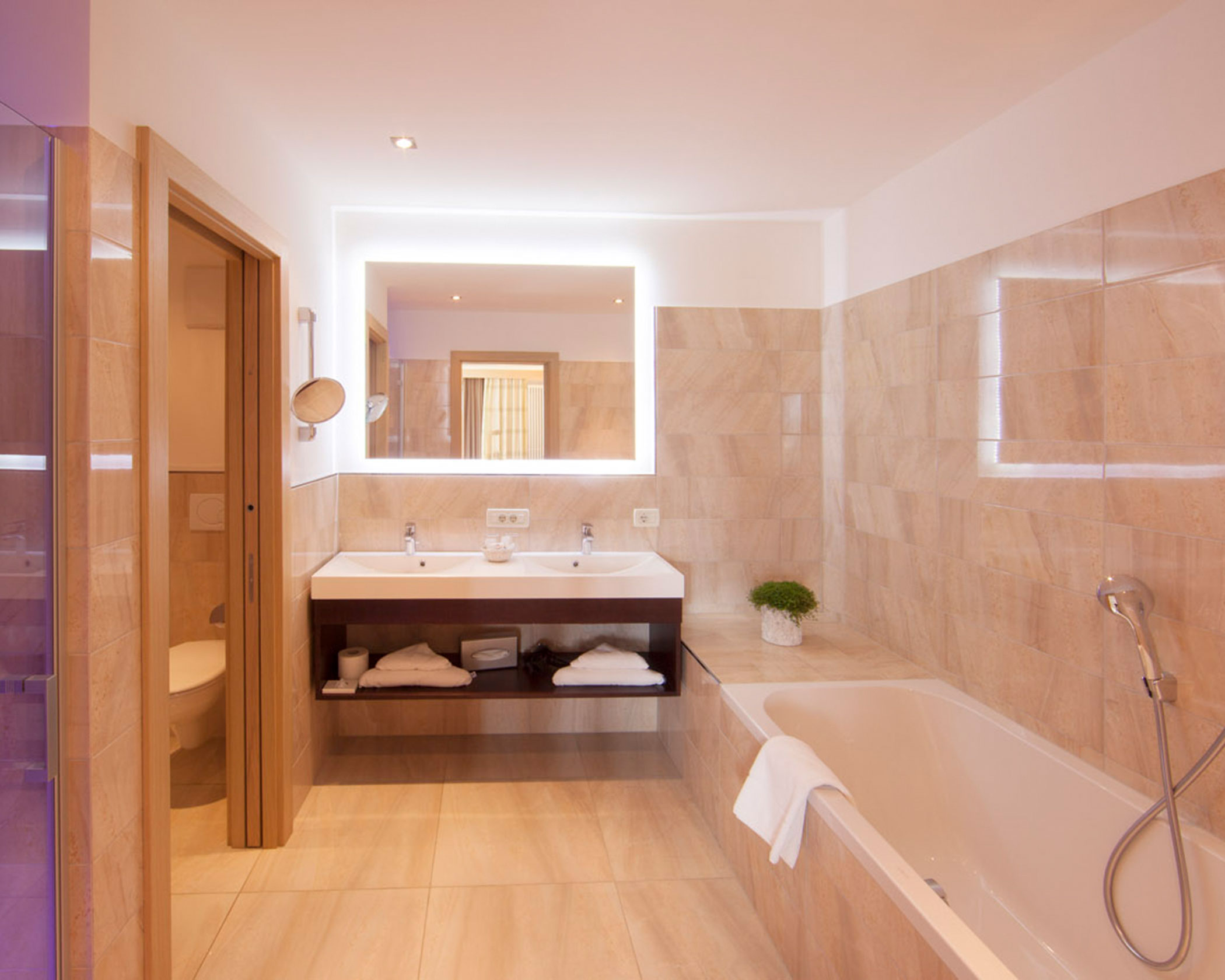 Modern bathroom with beige tiles, a double sink, large mirror, and a bathtub with a white towel draped over the edge. - Hotel Lagrein