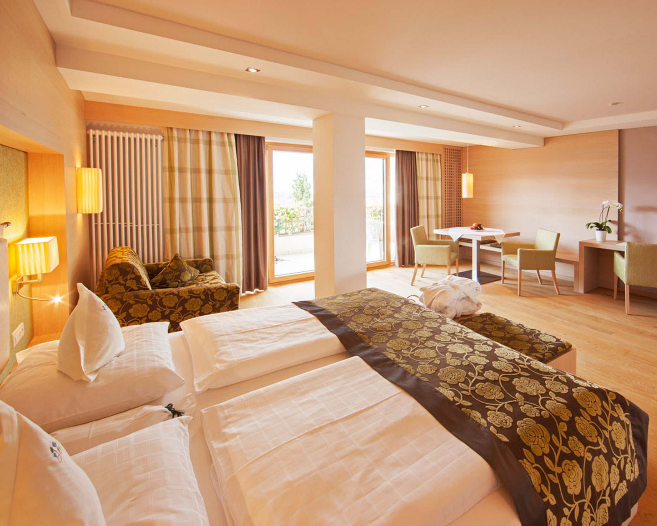 Spacious hotel room with a double bed, sofa, table with chairs, and large windows letting in natural light. - Hotel Lagrein