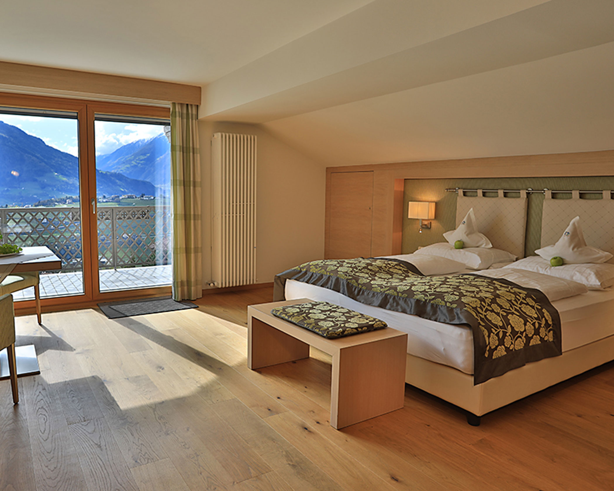 Sunlit hotel room with double bed, wooden floor, balcony, table, chairs, and mountain view through glass doors. - Hotel Lagrein