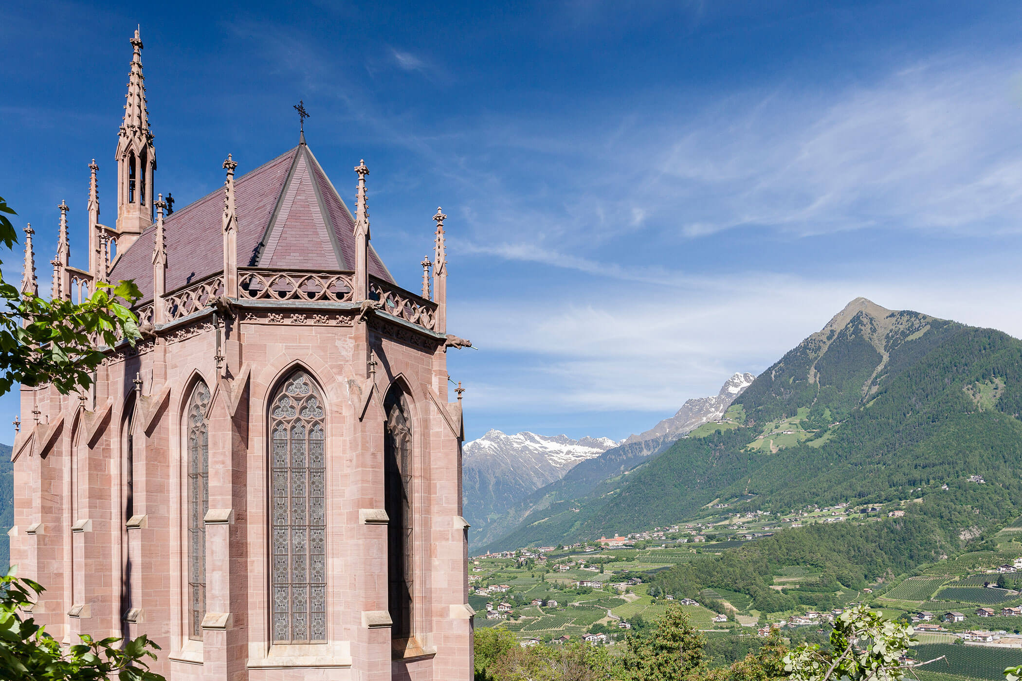 Gothic-style church tower with large arched windows, set against a backdrop of mountains and valley. - Hotel Lagrein