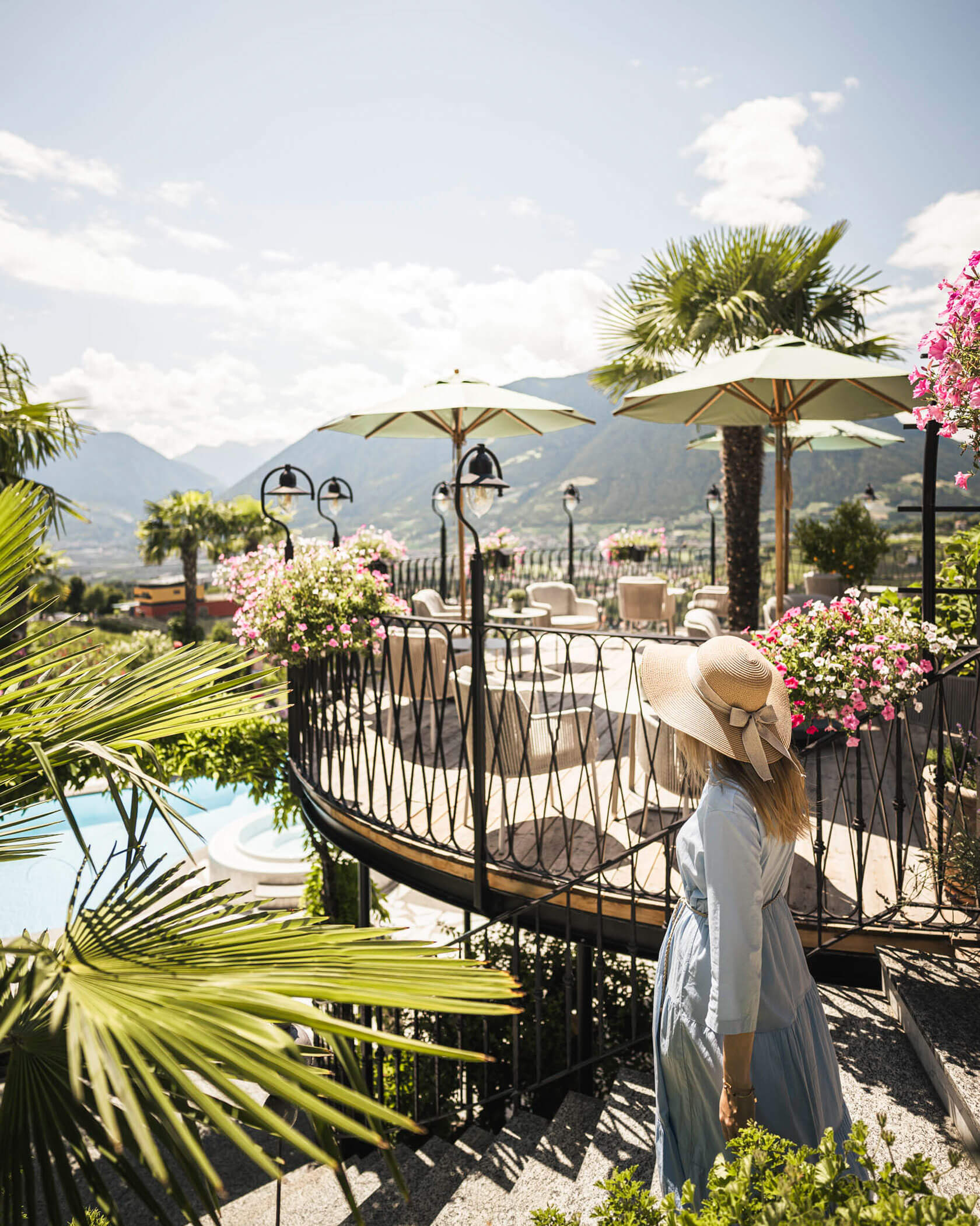 A woman in a sunhat walks by a terrace with flowers, umbrellas, and mountain views on a sunny day. - Hotel Lagrein