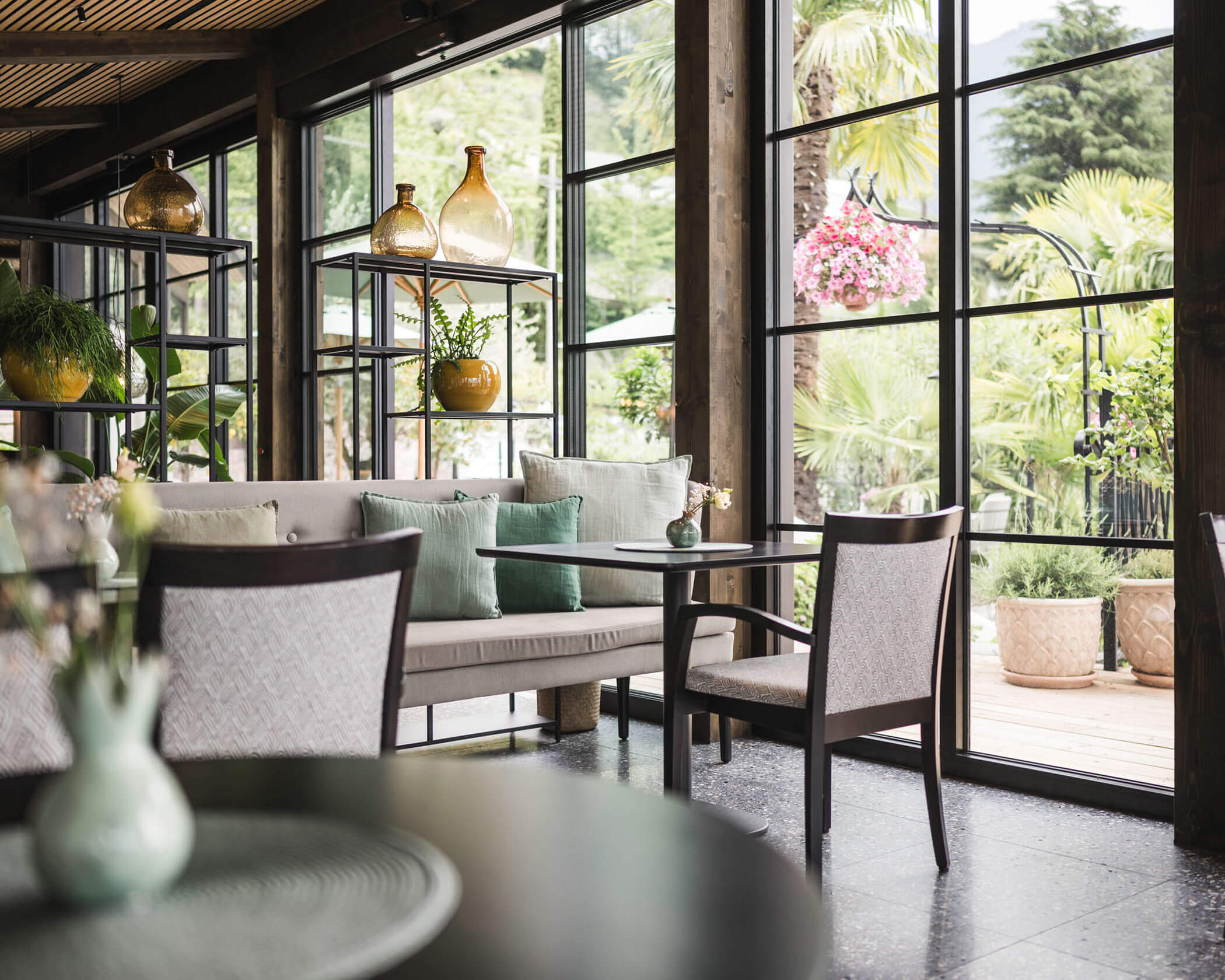 Modern cafe with large windows, elegant decor, light cushions, and lush greenery visible outside. - Hotel Lagrein