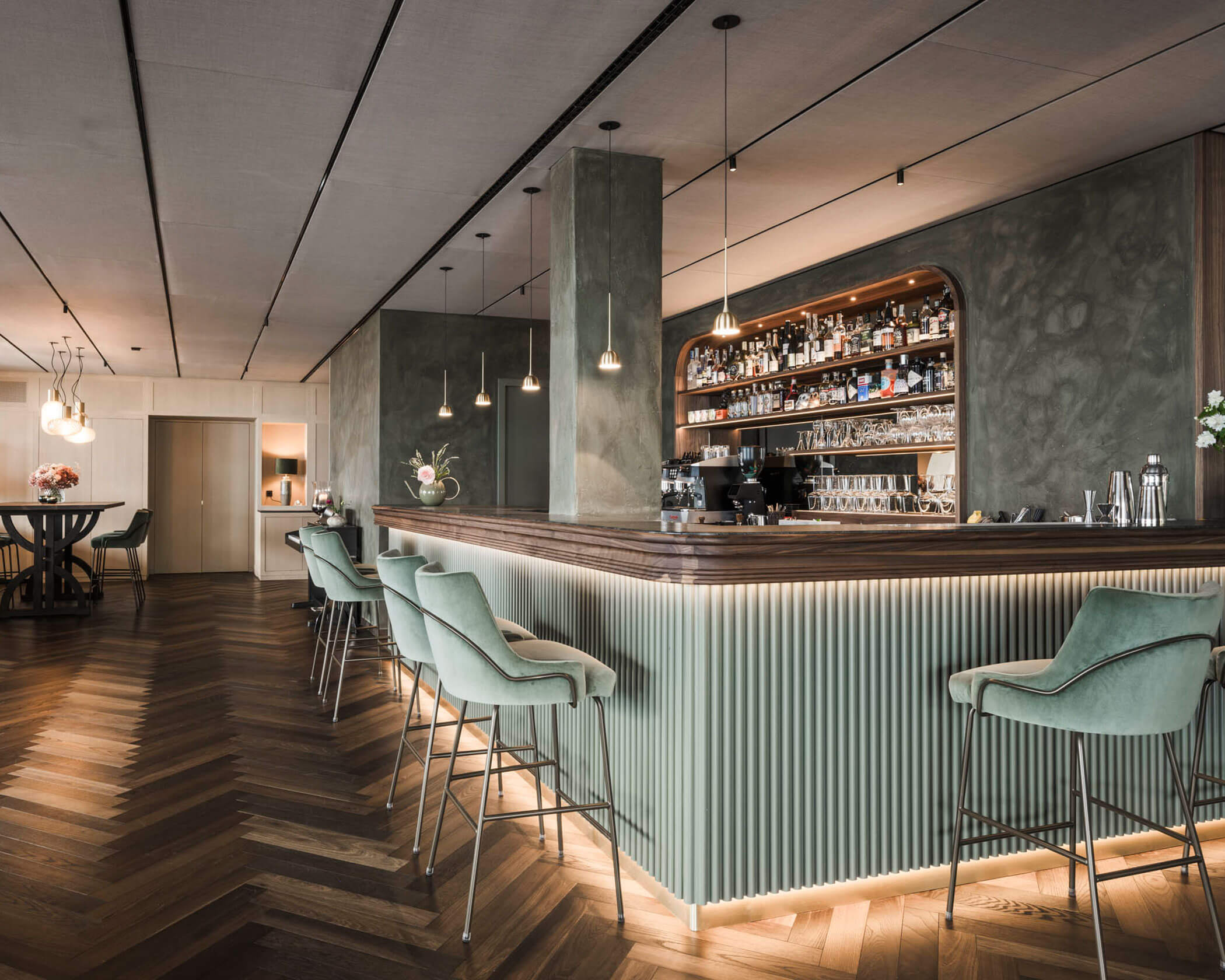 Modern bar with mint green stools, wooden floors, backlit counter, and shelves stocked with bottles and glasses. - Hotel Lagrein