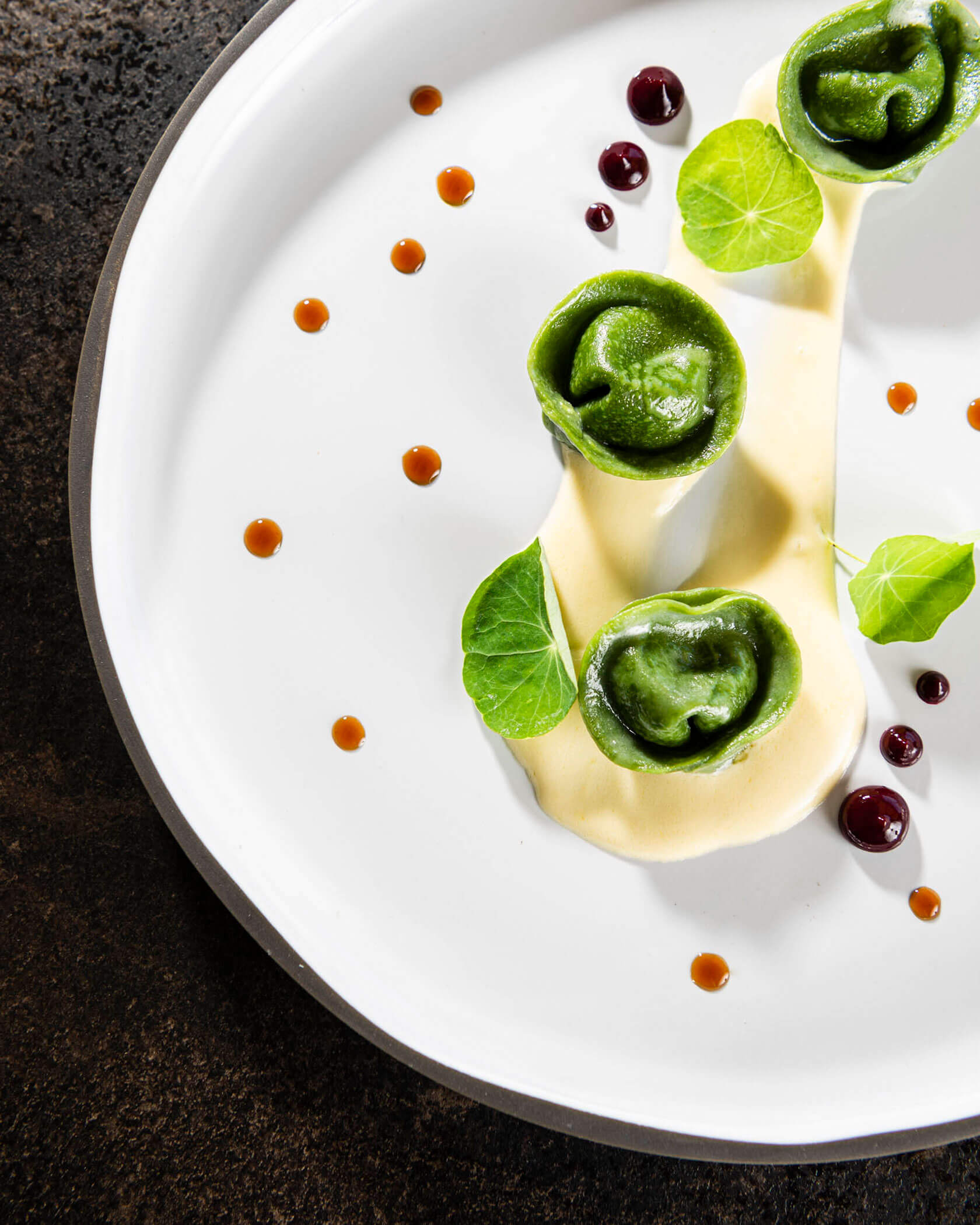 Three green tortellini on a white plate with creamy sauce, garnish, and dots of red and brown sauce. - Hotel Lagrein