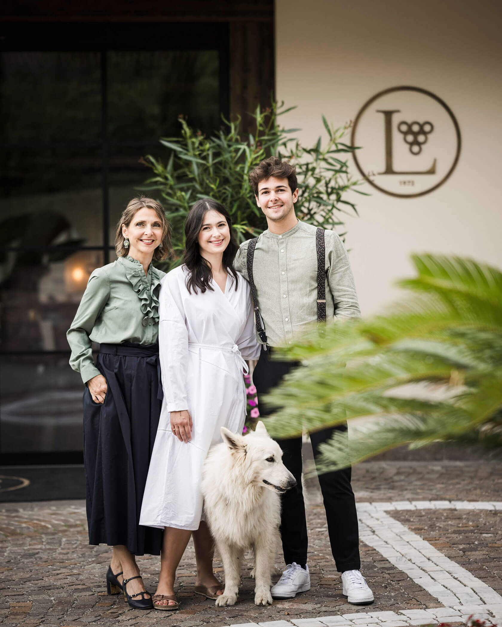 Three people and a white dog stand smiling outdoors in front of a building with a grape-themed logo. - Hotel Lagrein