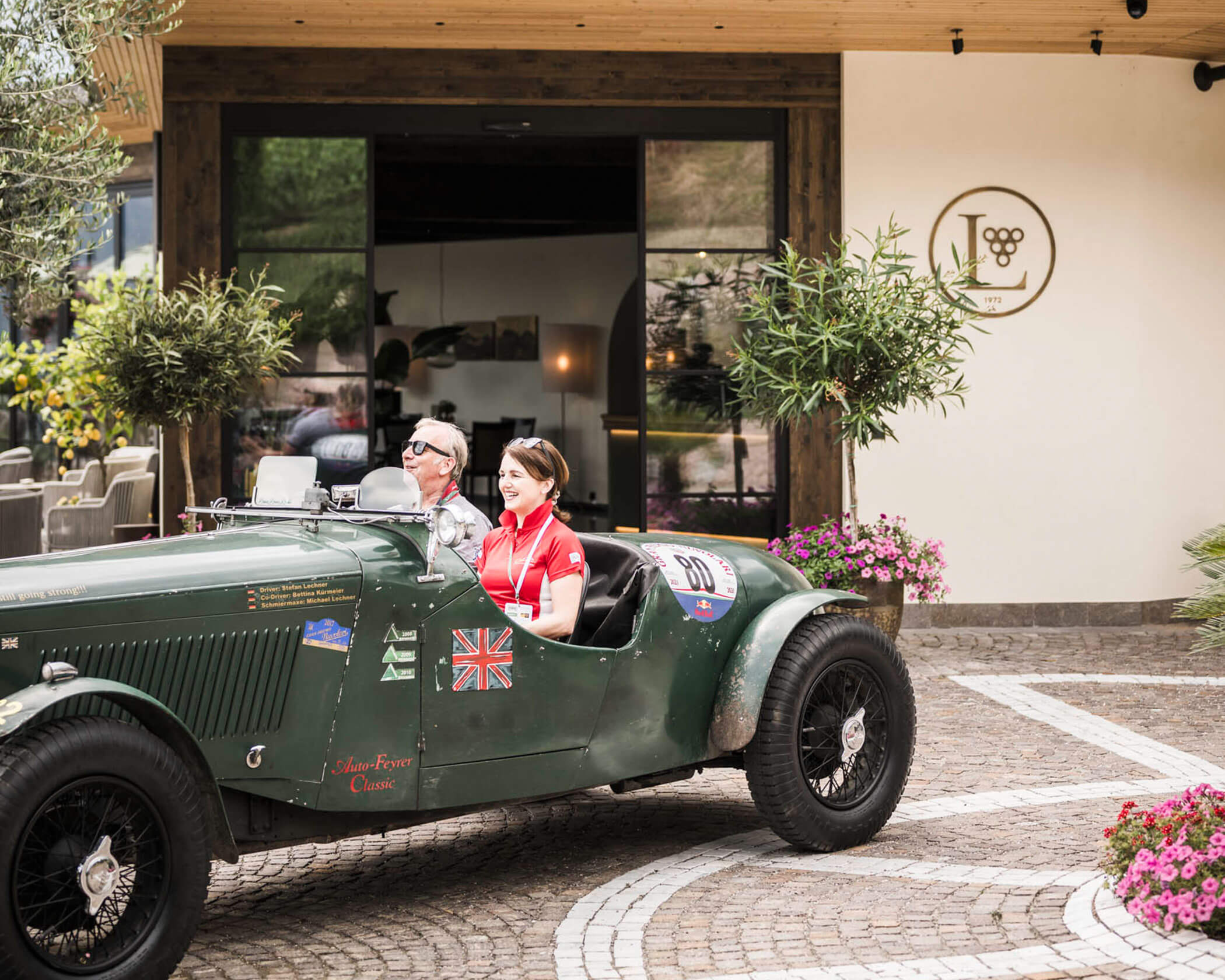 Two people drive a vintage green car past a building with a JL logo and potted plants outside the entrance. - Hotel Lagrein