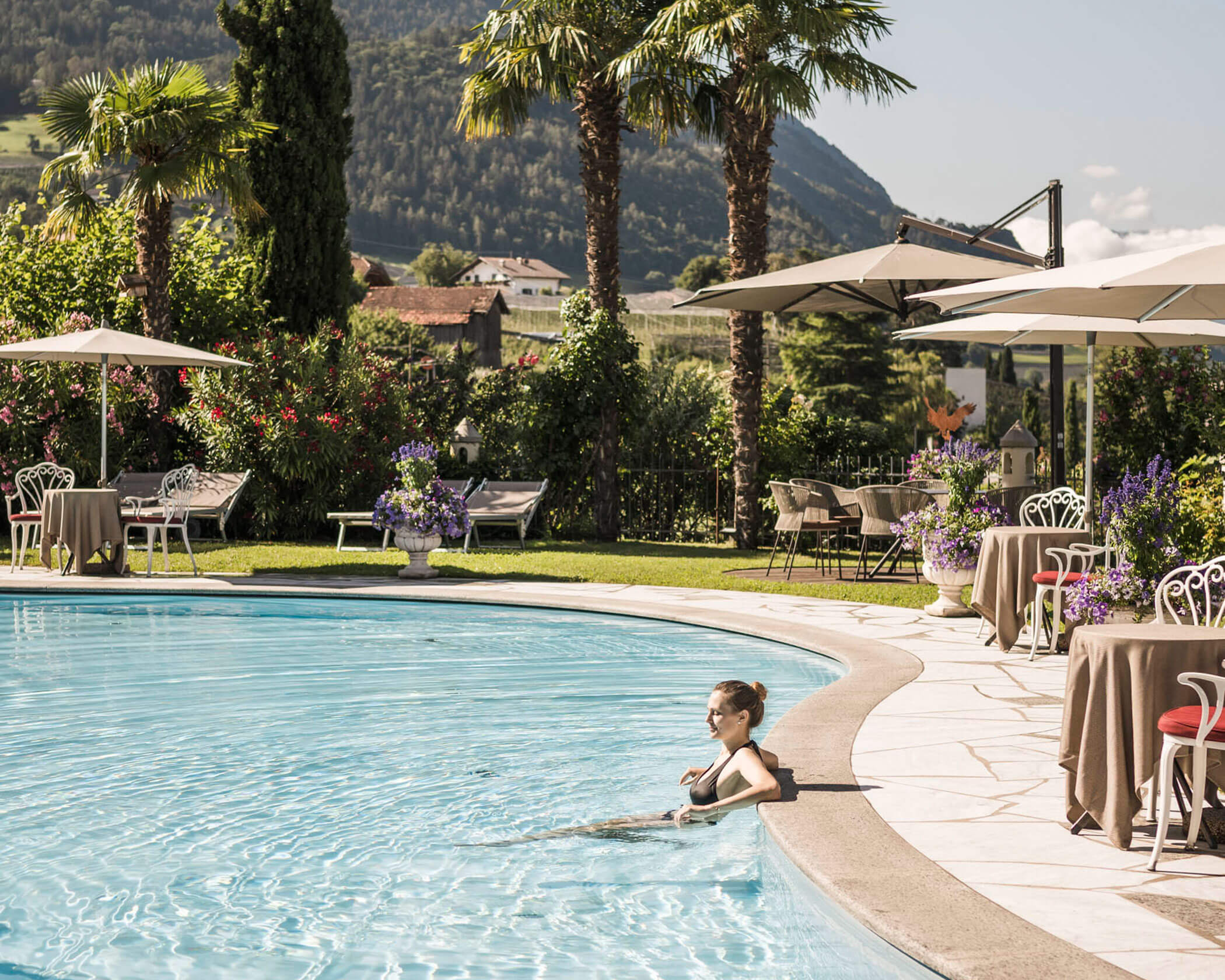 Woman relaxing in a pool, surrounded by palm trees, tables, and mountains in the background. - Hotel Lagrein