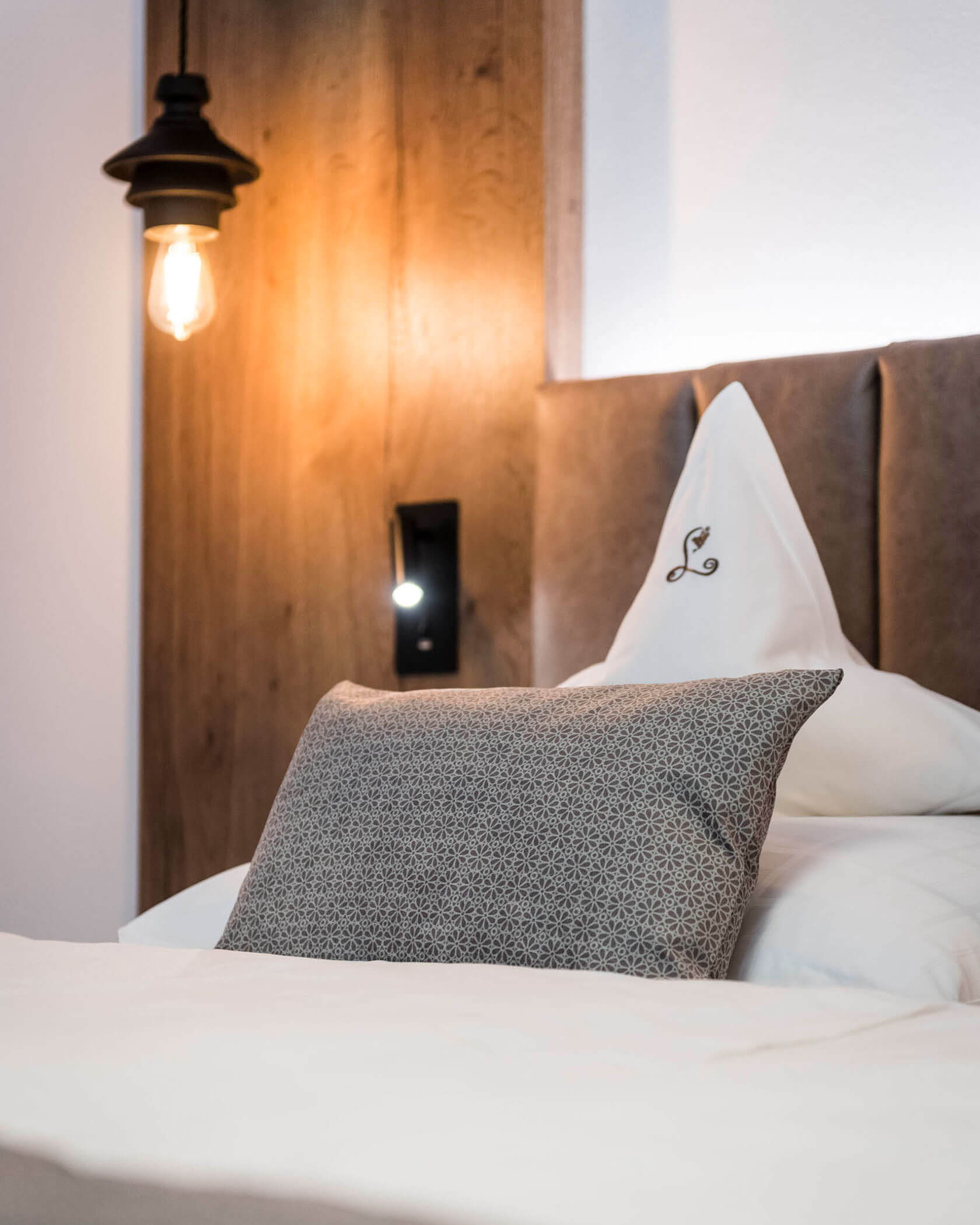A neatly made bed with patterned pillow, white linens, and a lit bulb on a wooden headboard. - Hotel Lagrein