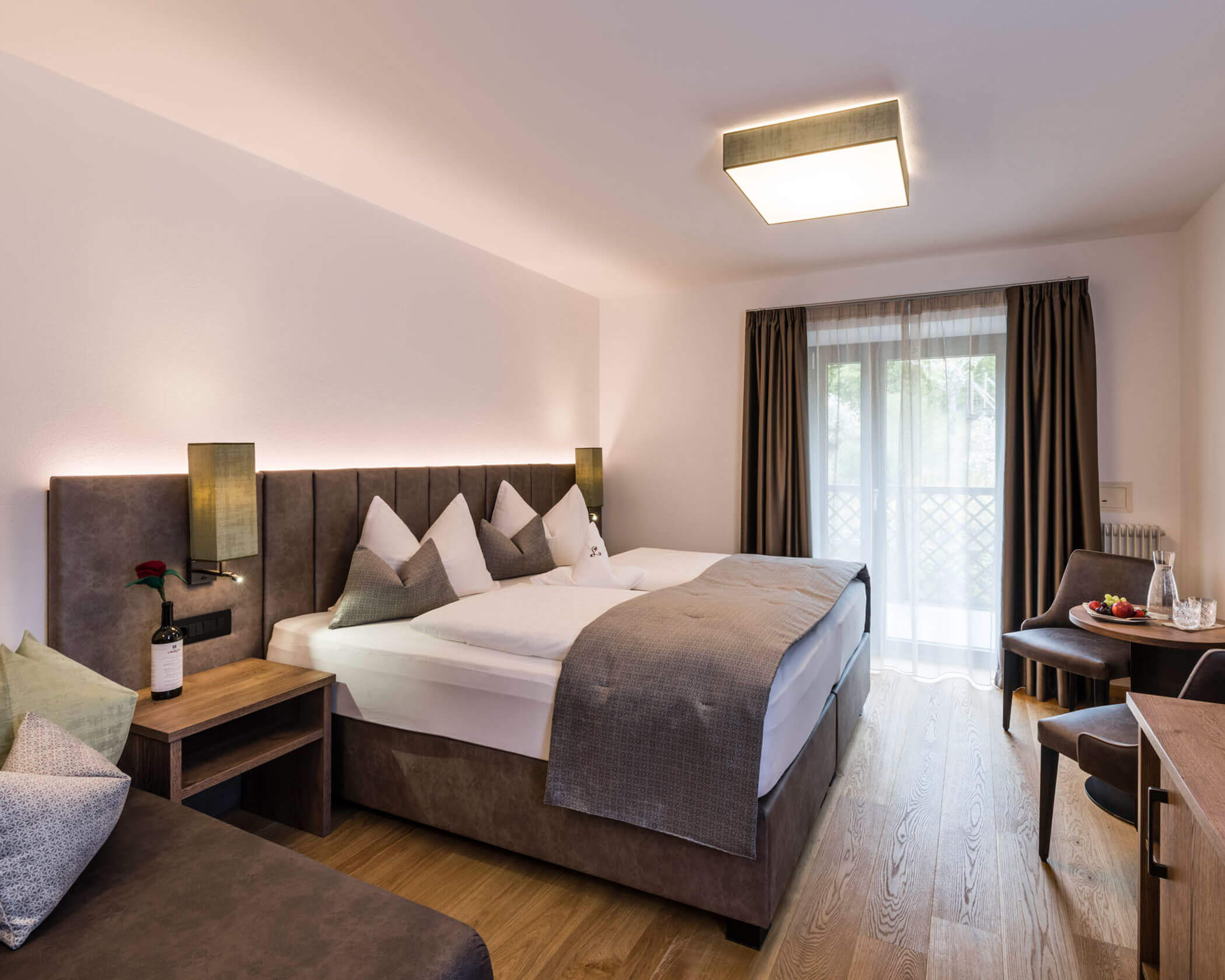 Modern hotel room with a double bed, sofa, table, chairs, and a balcony with curtains. - Hotel Lagrein