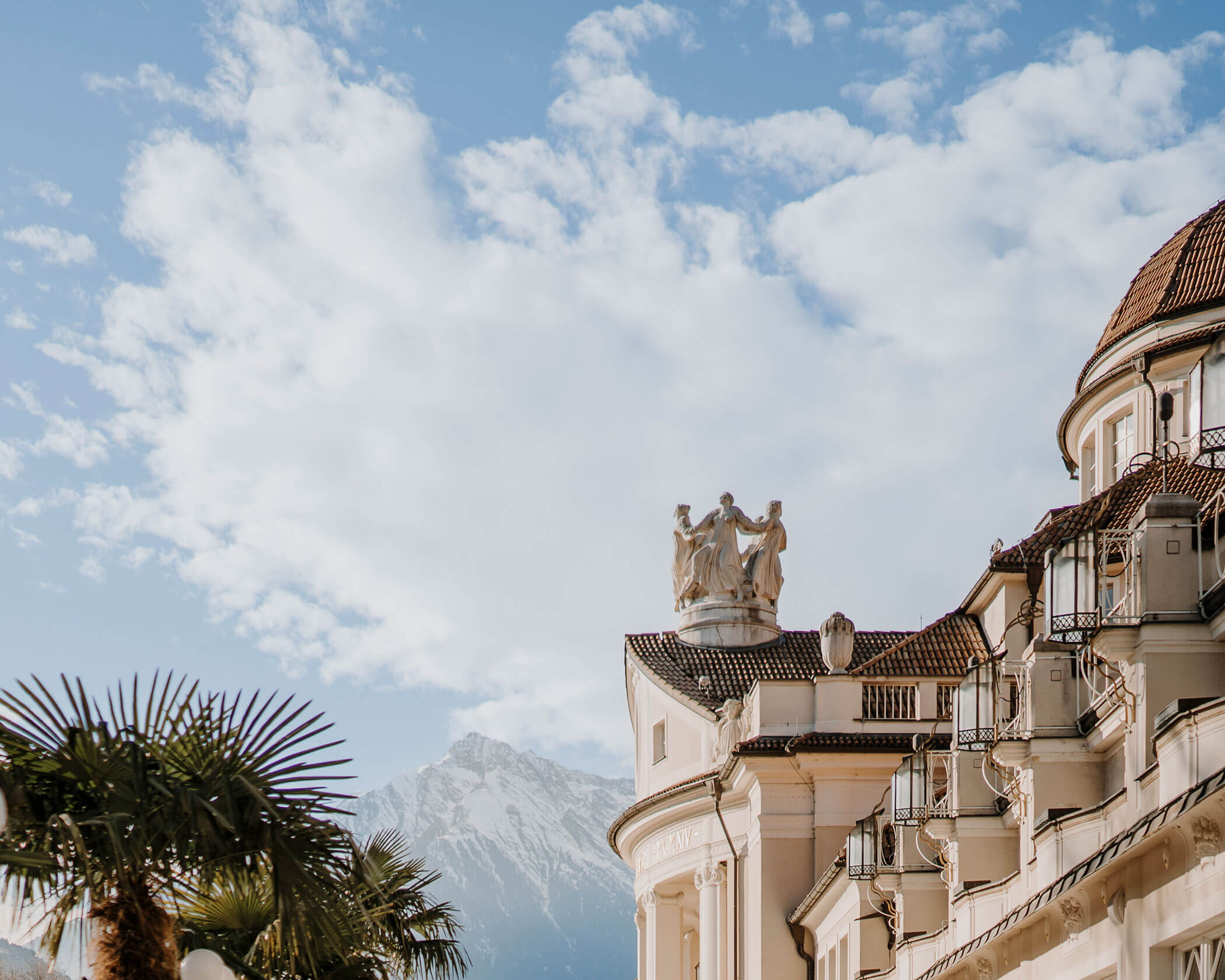 Ornate building with statues, palm trees, and mountains in the background under a partly cloudy sky. - Hotel Lagrein