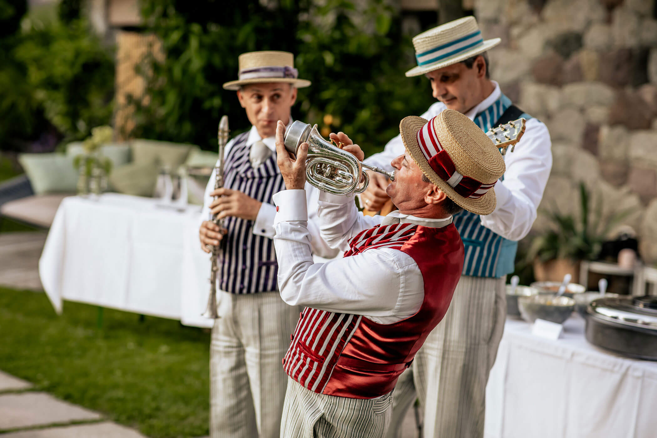 Three musicians in vintage attire perform outdoors; one plays a horn energetically in the foreground. - Hotel Lagrein