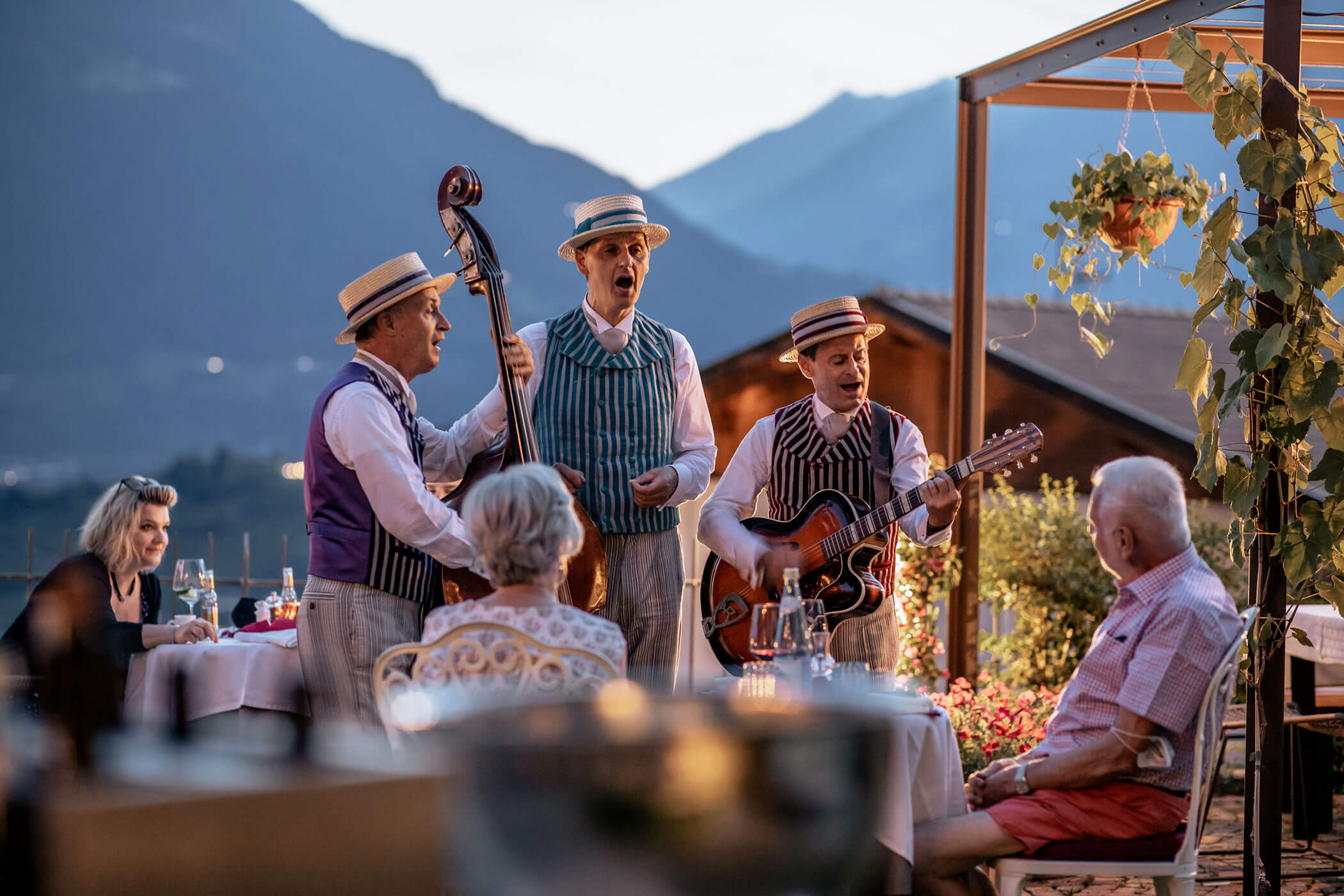 Three musicians in vintage attire perform outdoors for seated diners with mountains in the background. - Hotel Lagrein