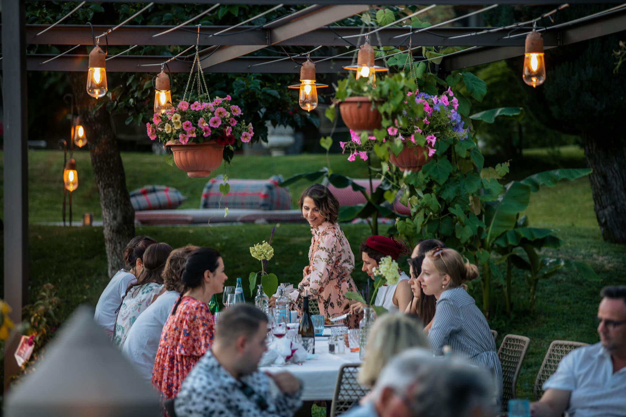 People dining outdoors at a garden table under string lights and hanging flower baskets on a summer evening. - Hotel Lagrein