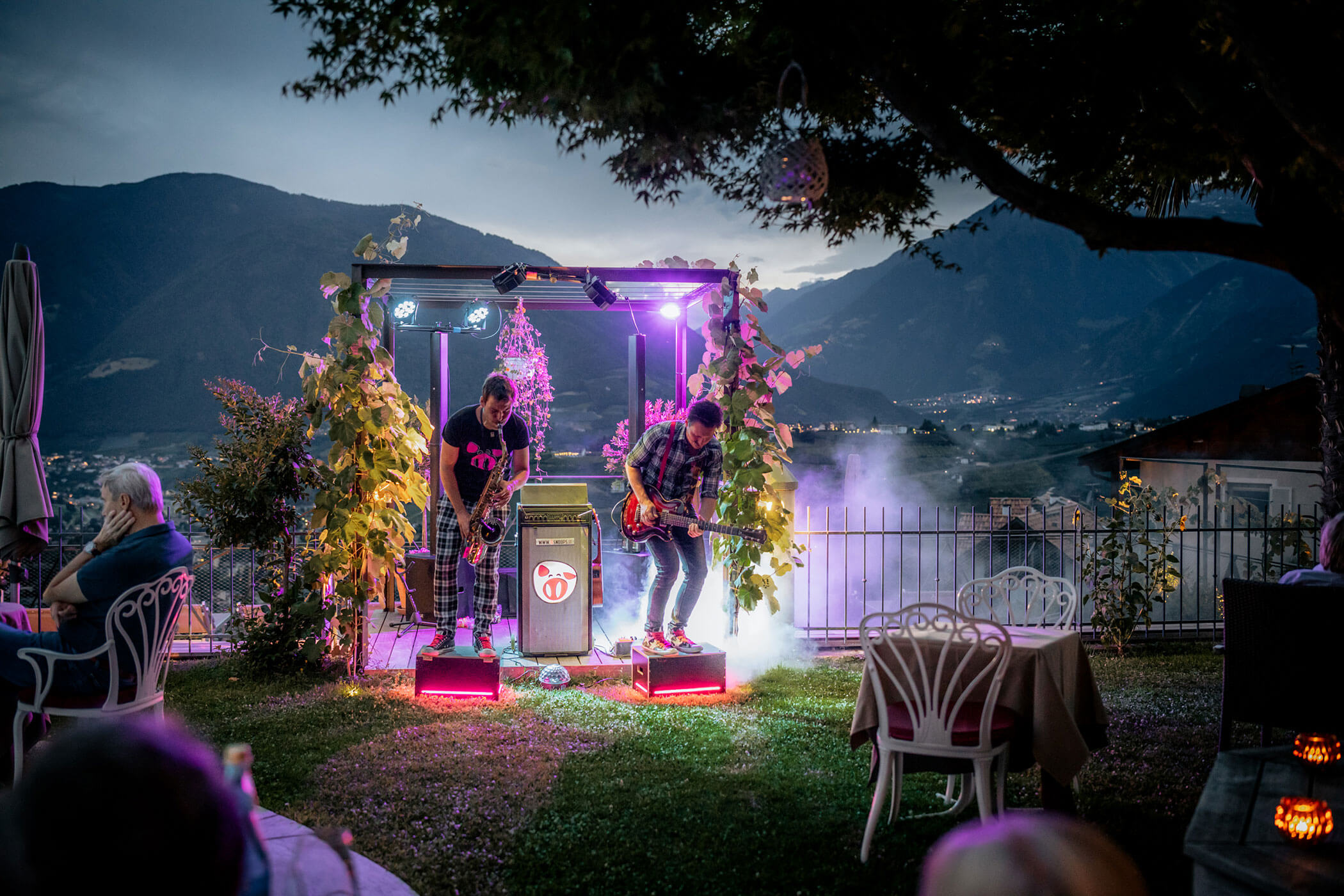 Two musicians perform outdoors on lit platforms with mountains in the background and smoke effects. - Hotel Lagrein