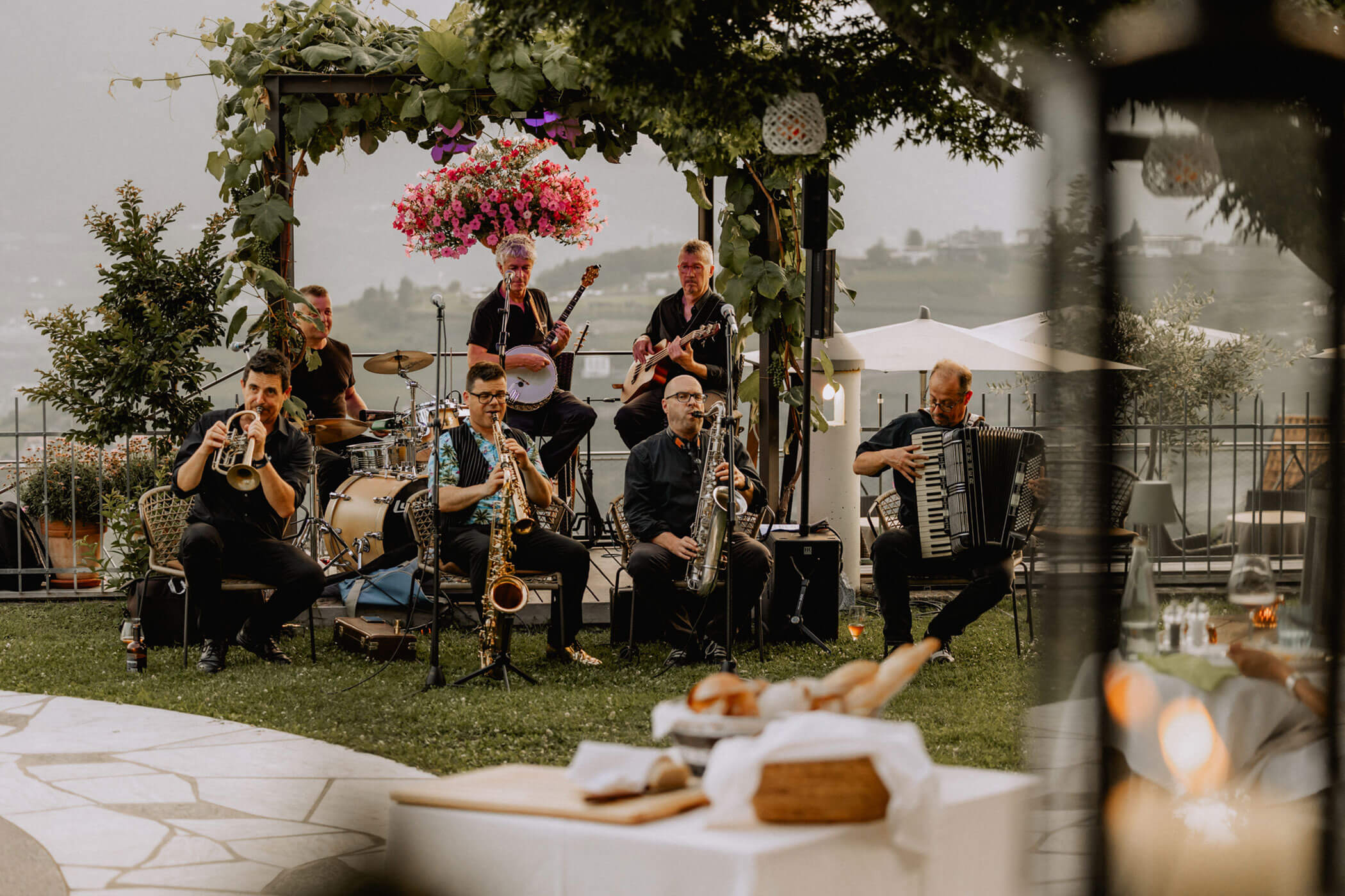 A jazz band performs outdoors on a lawn, surrounded by trees and flowers, with food on a nearby table. - Hotel Lagrein