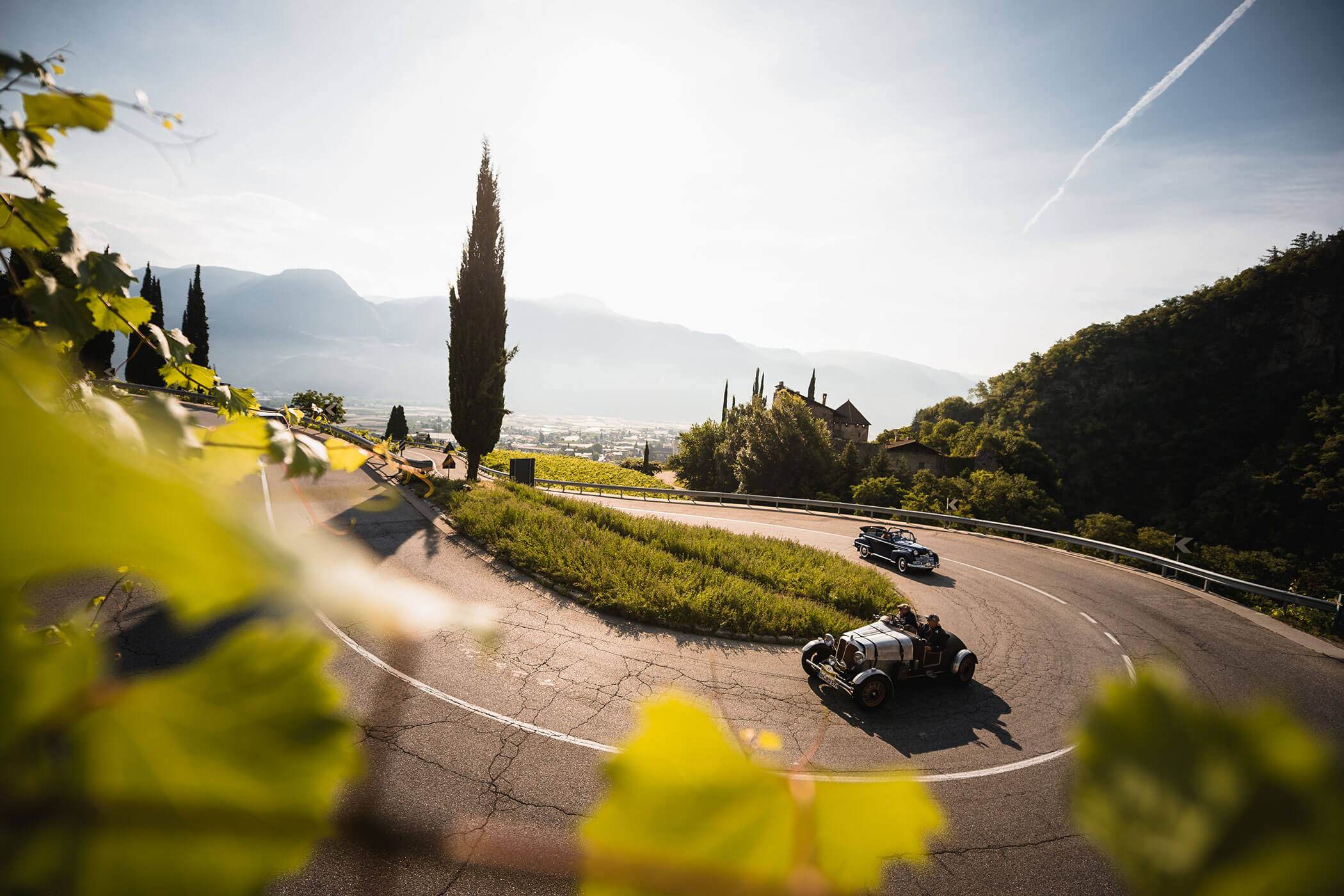 Two vintage cars drive along a winding mountain road with greenery and a sunlit valley in the background. - Hotel Lagrein