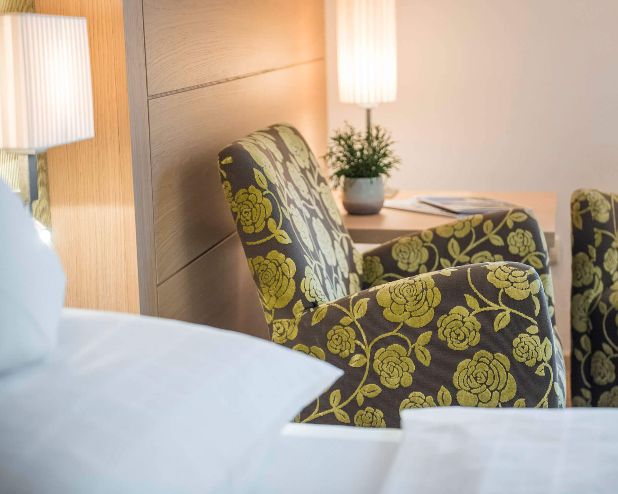 Floral armchair by a desk with a potted plant, beside a bed and softly lit lamps in a cozy room. - Hotel Lagrein