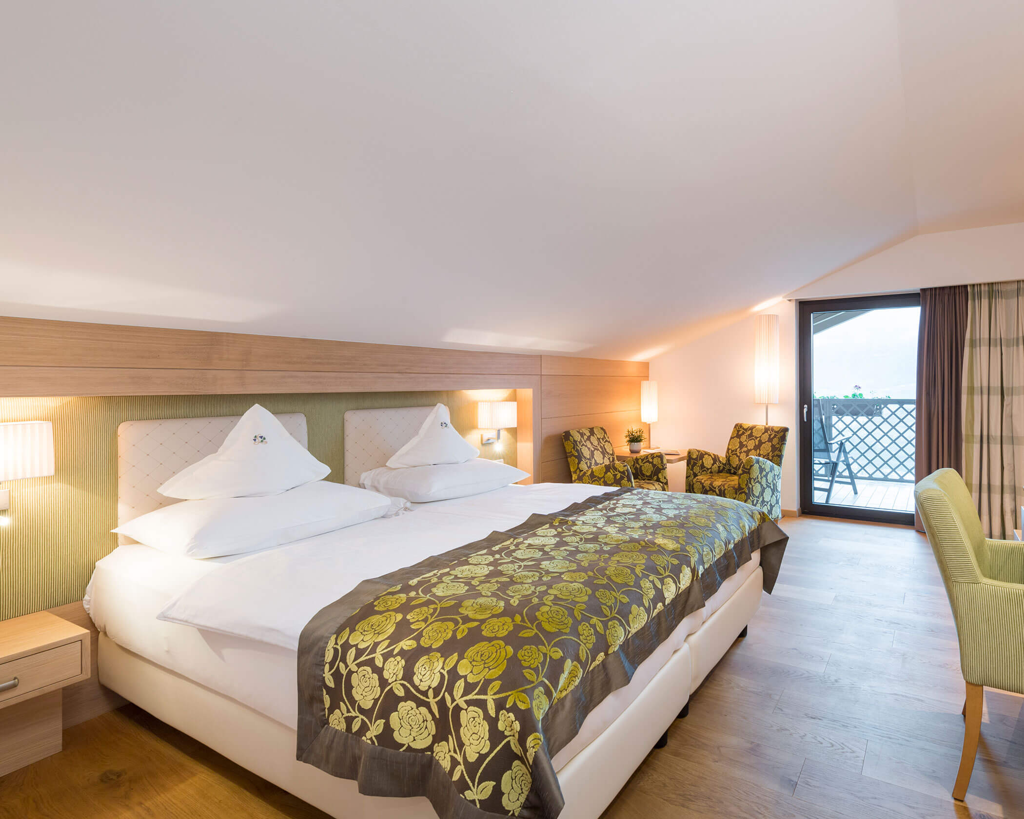 Bright hotel room with a double bed, floral decor, chairs, and a balcony door letting in natural light. - Hotel Lagrein