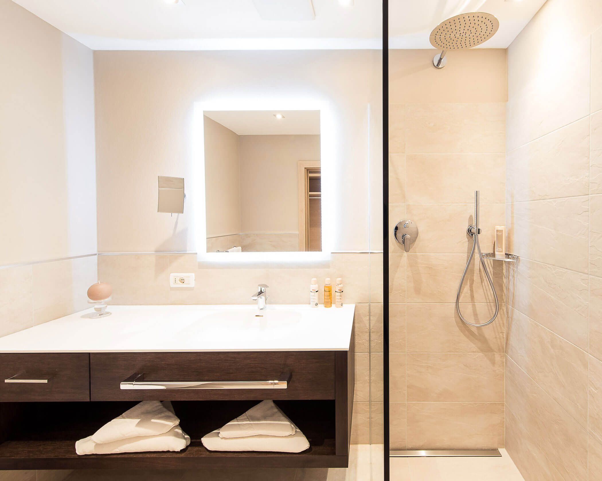 Modern bathroom with a lit mirror, sink, towels, and a walk-in shower with a rain showerhead. - Hotel Lagrein