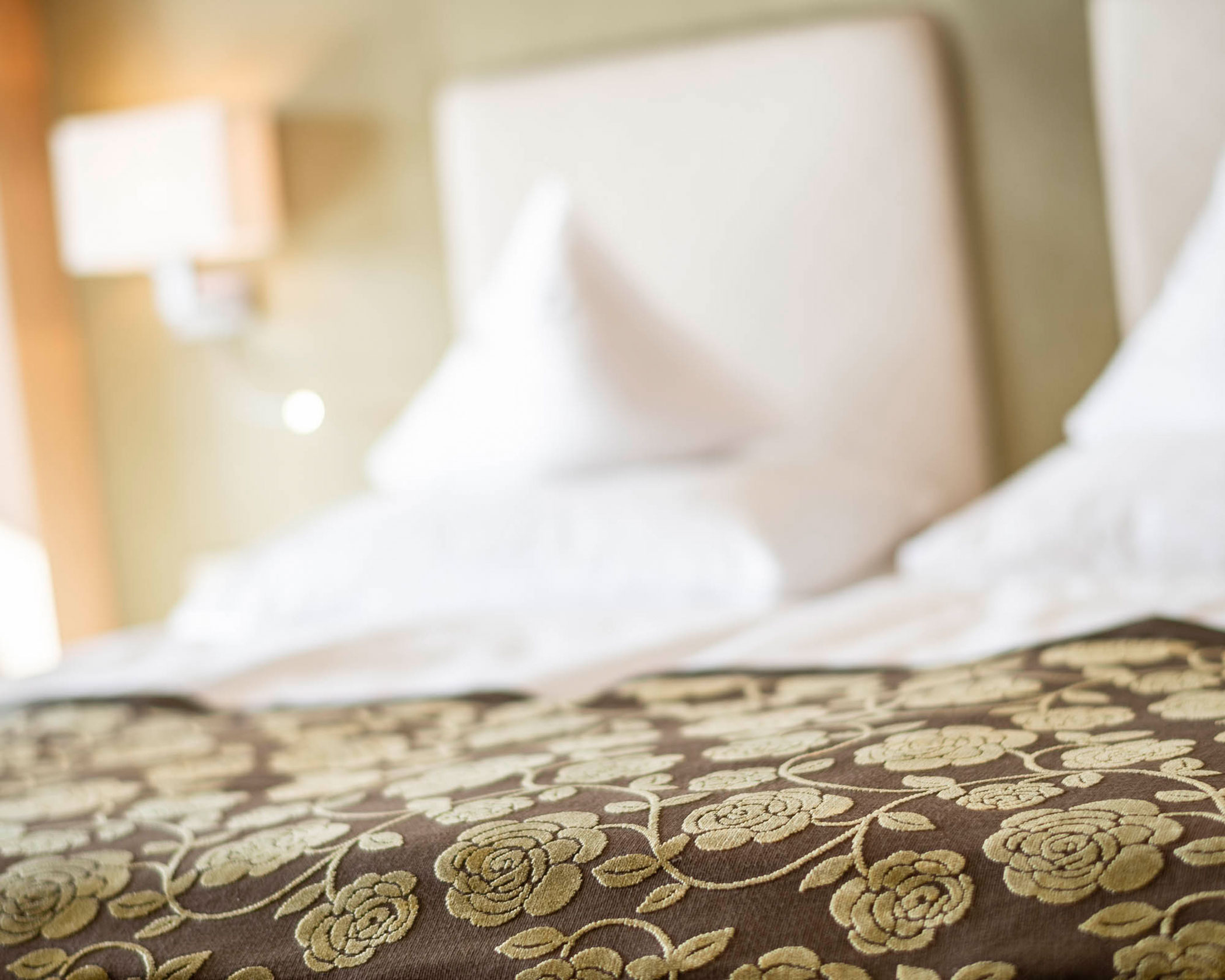 Close-up of a bed with white pillows and a brown floral patterned bedspread in a softly lit room. - Hotel Lagrein