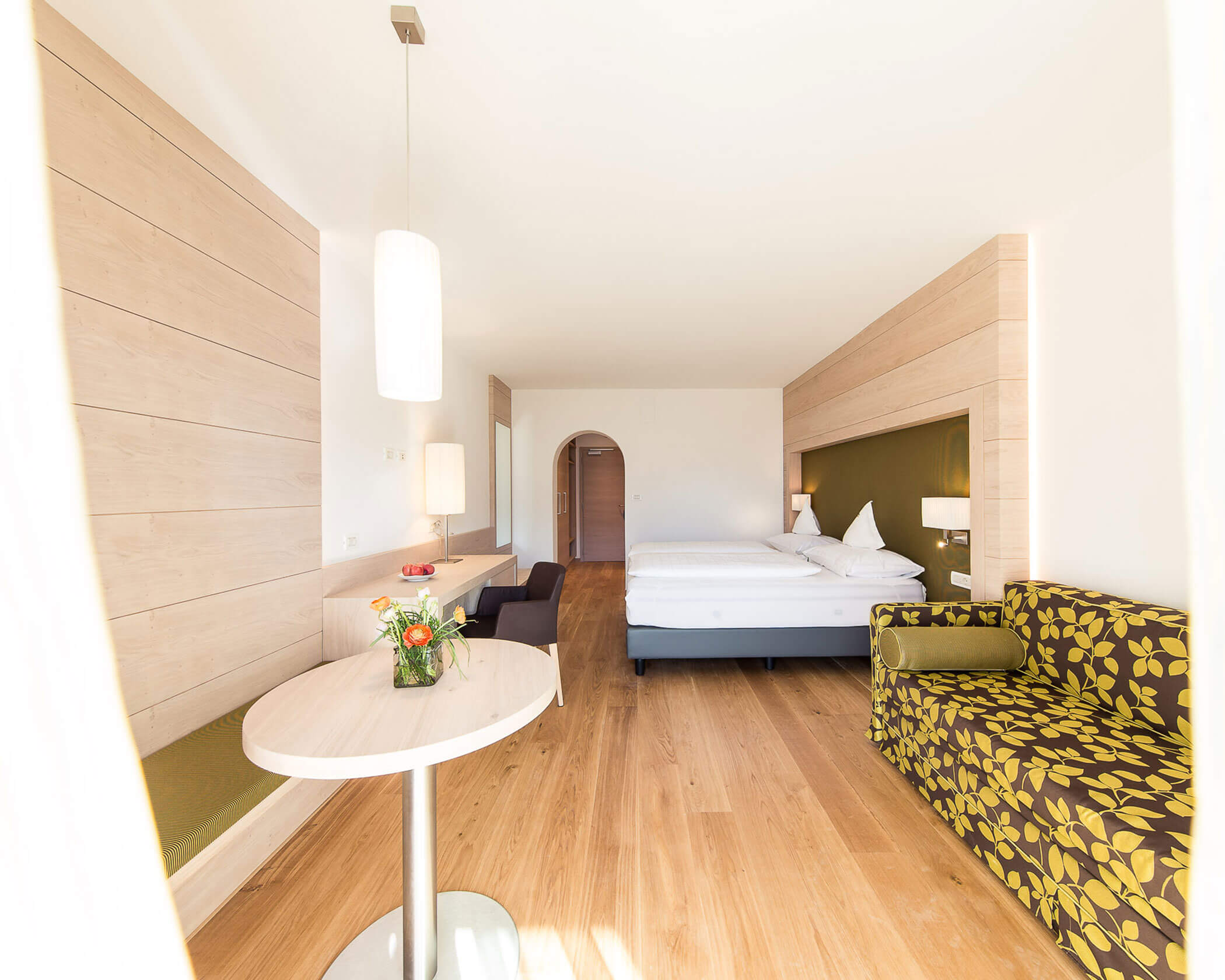 Bright hotel room with a double bed, green-patterned sofa, and a small round table with a flower vase. - Hotel Lagrein