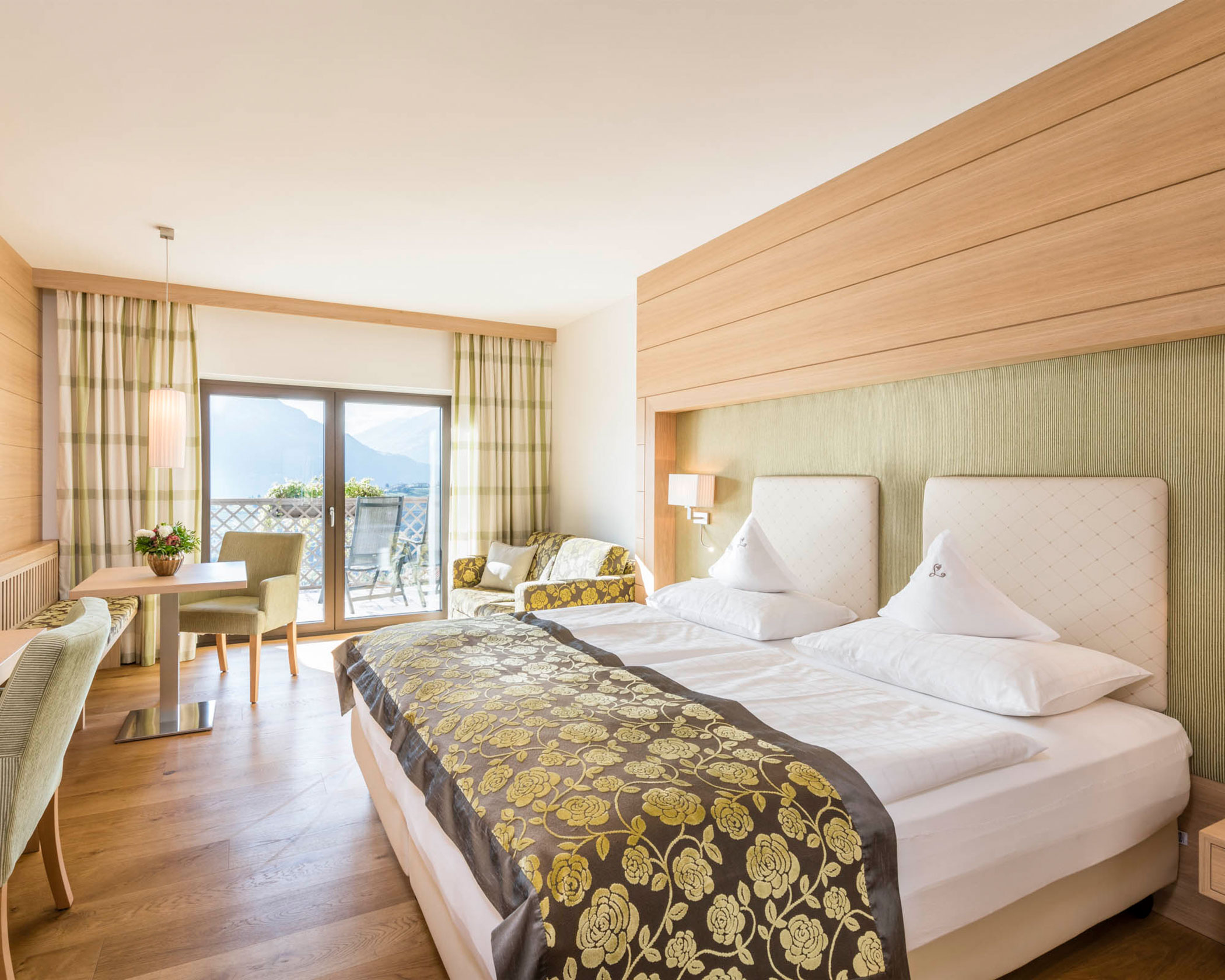 Bright hotel room with double bed, floral bedding, seating area, desk, and balcony overlooking mountains. - Hotel Lagrein