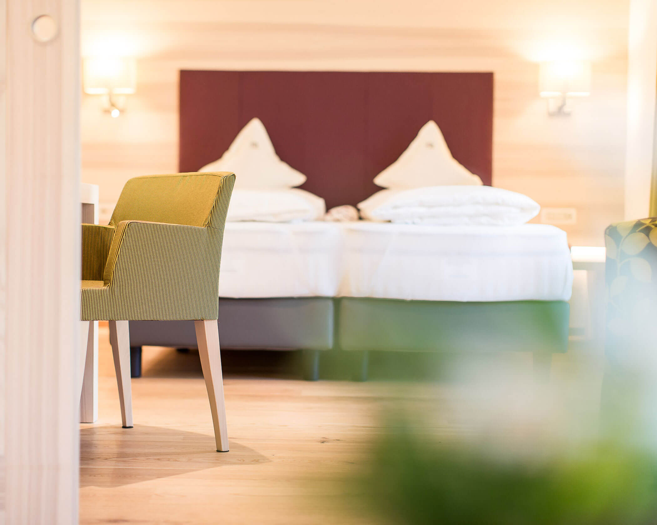 A modern hotel room with twin beds, green chair, and soft lighting on beige walls. - Hotel Lagrein