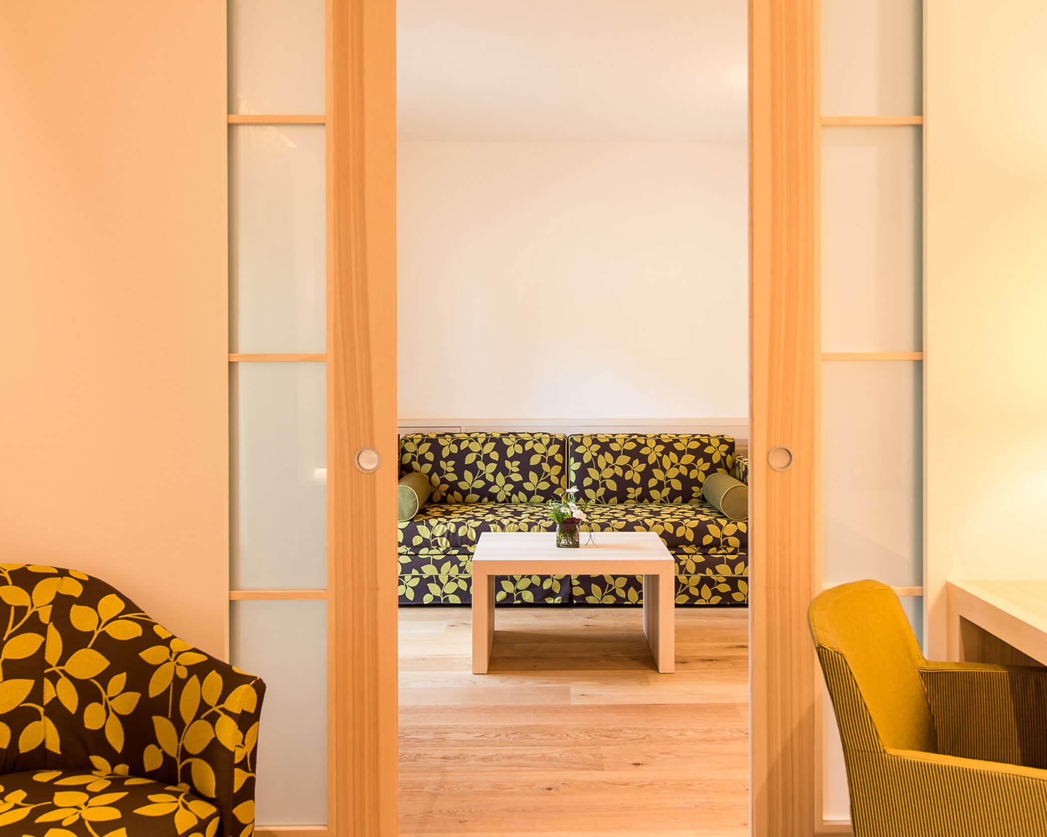 Room with a floral-patterned sofa and armchair, sliding wooden doors, and a small table with a plant. - Hotel Lagrein
